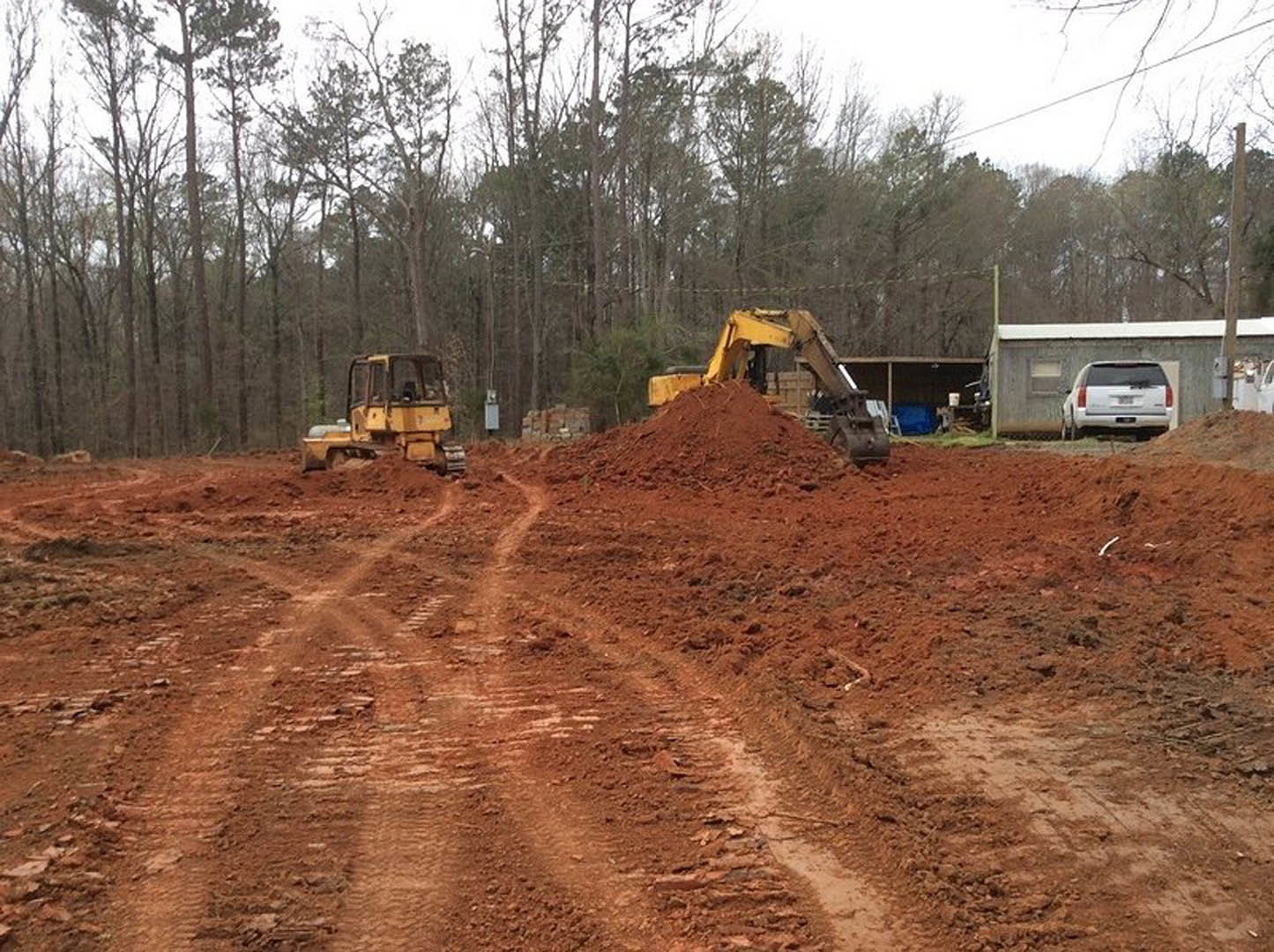 Yellow excavator digging into a large dirt pile on a residential construction site, with tire tracks visible on the dirt road and a white car parked nearby; trees and sky in the