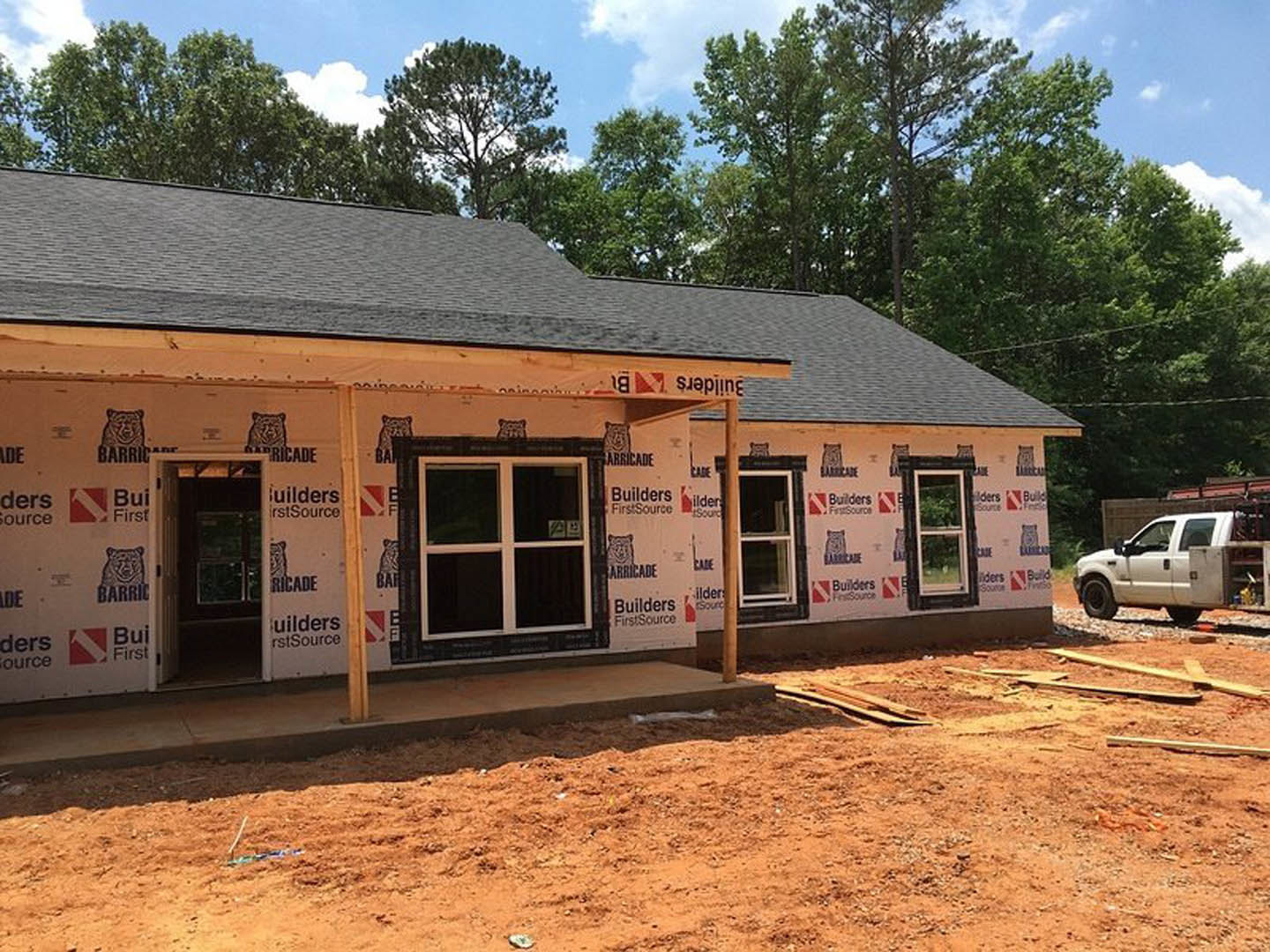 Partially built house with exposed framing, white truck parked in driveway, white-framed windows, porch area, green plant visible through window, cloudy sky overhead