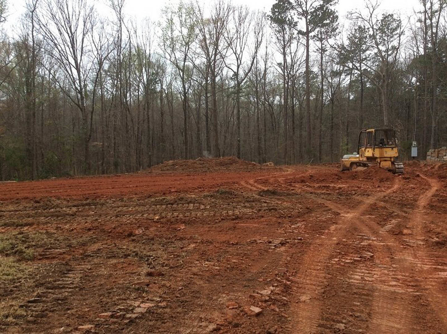Yellow bulldozer parked on a dirt field with tire tracks, surrounded by trees and a pile of bricks under a clear sky