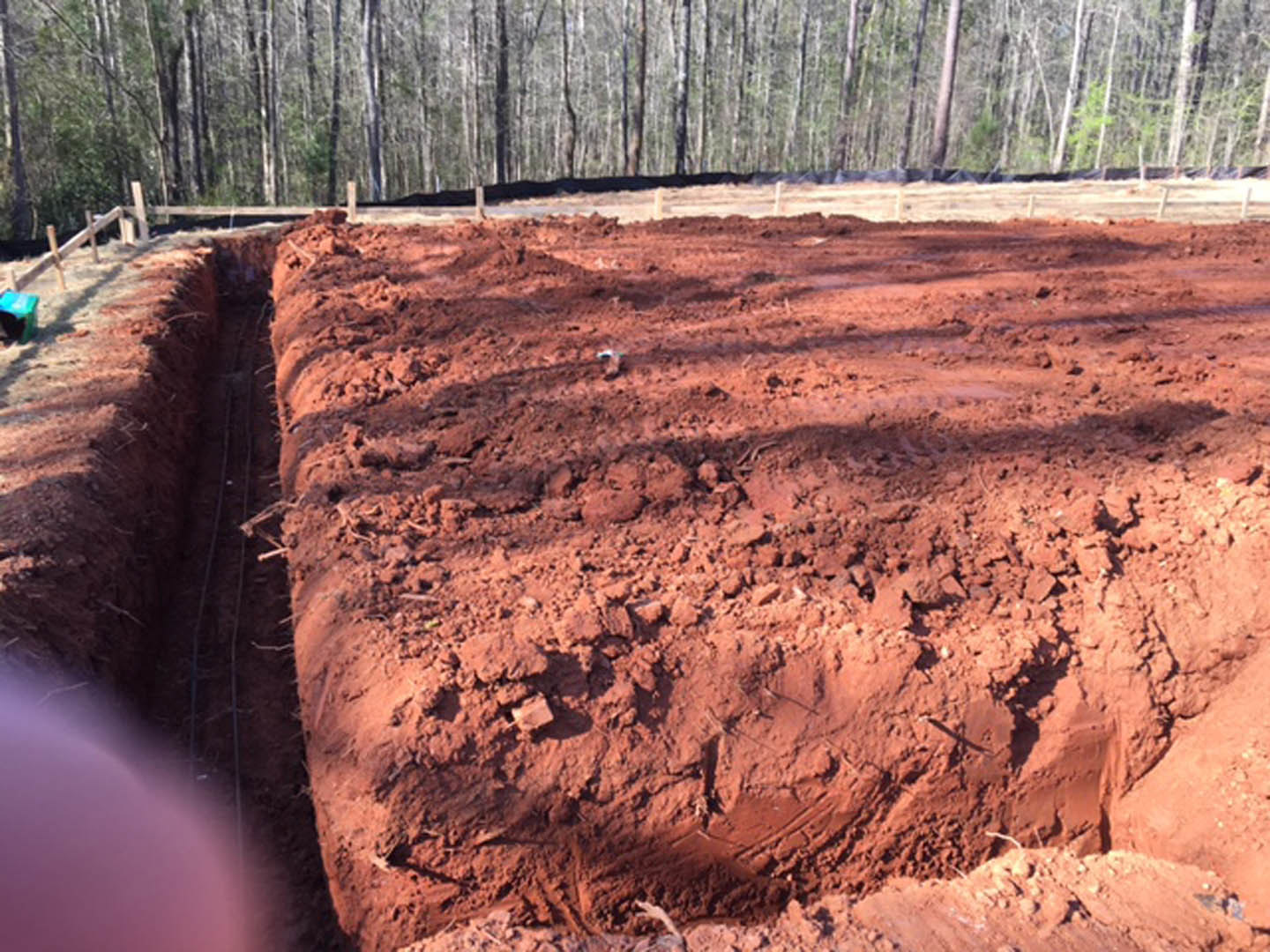 Red dirt field with a deep trench in the foreground, bordered by trees and plants in the background