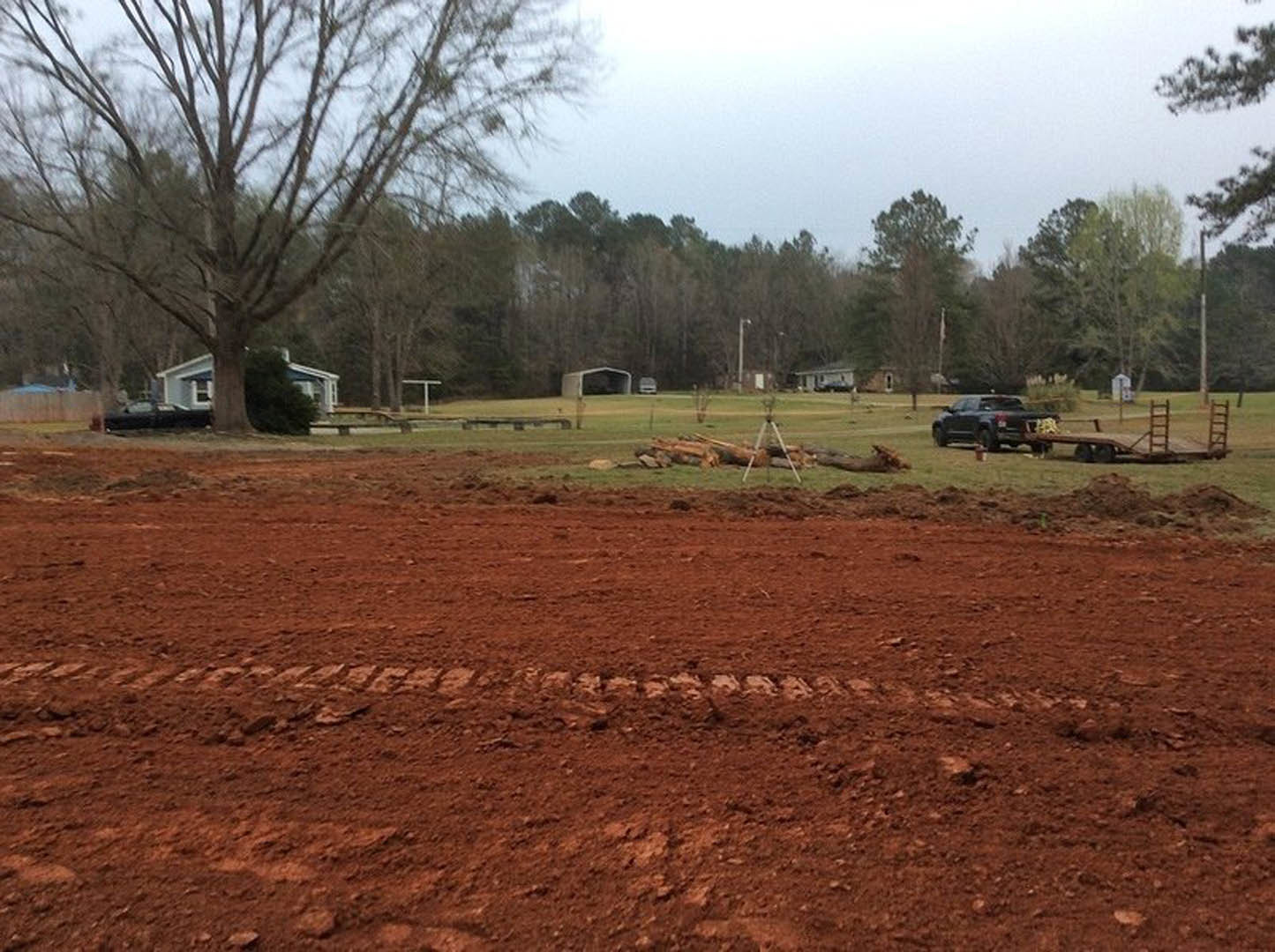 Modern house with light siding and large windows set behind a leafless tree, surrounded by a dirt field with visible tire tracks and sparse grass.