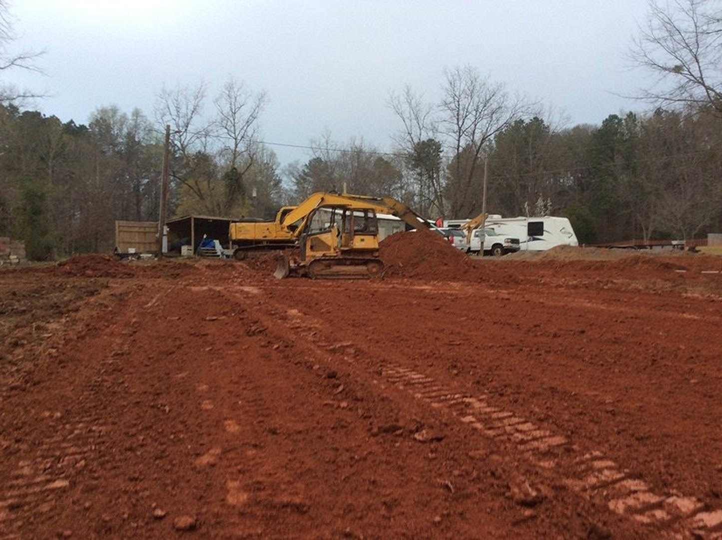 Yellow bulldozer with large wheels digging into brown soil on a residential construction site, surrounded by trees and under a cloudy sky