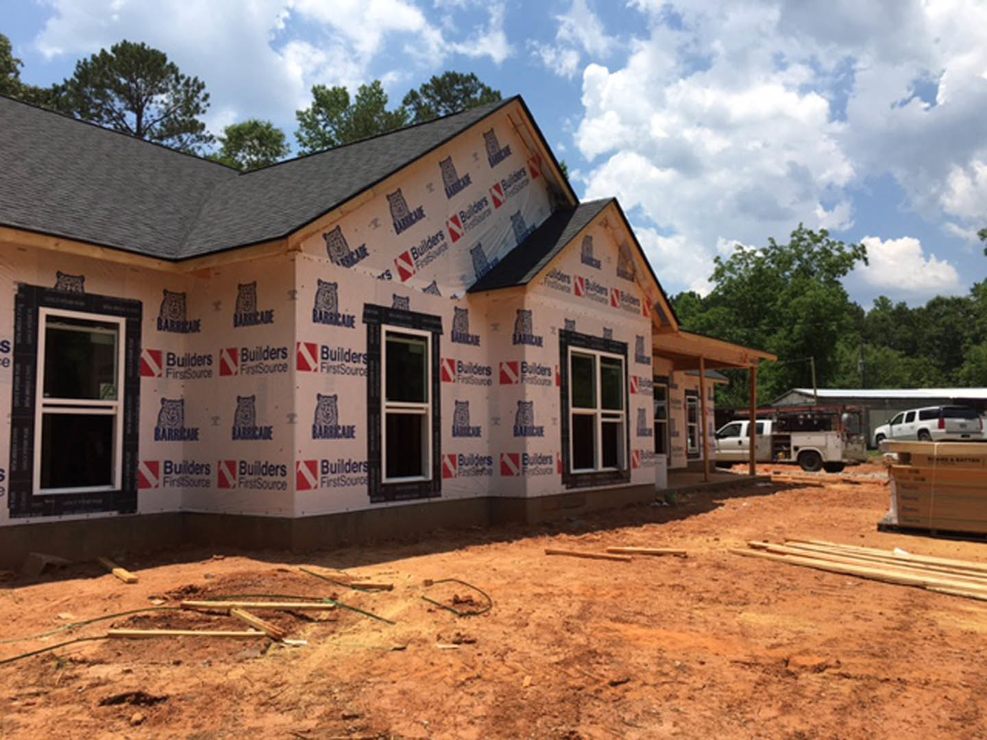 Partially built house with exposed framing and sheathing, surrounded by red dirt under a clear blue sky, white-framed windows visible on exterior wall