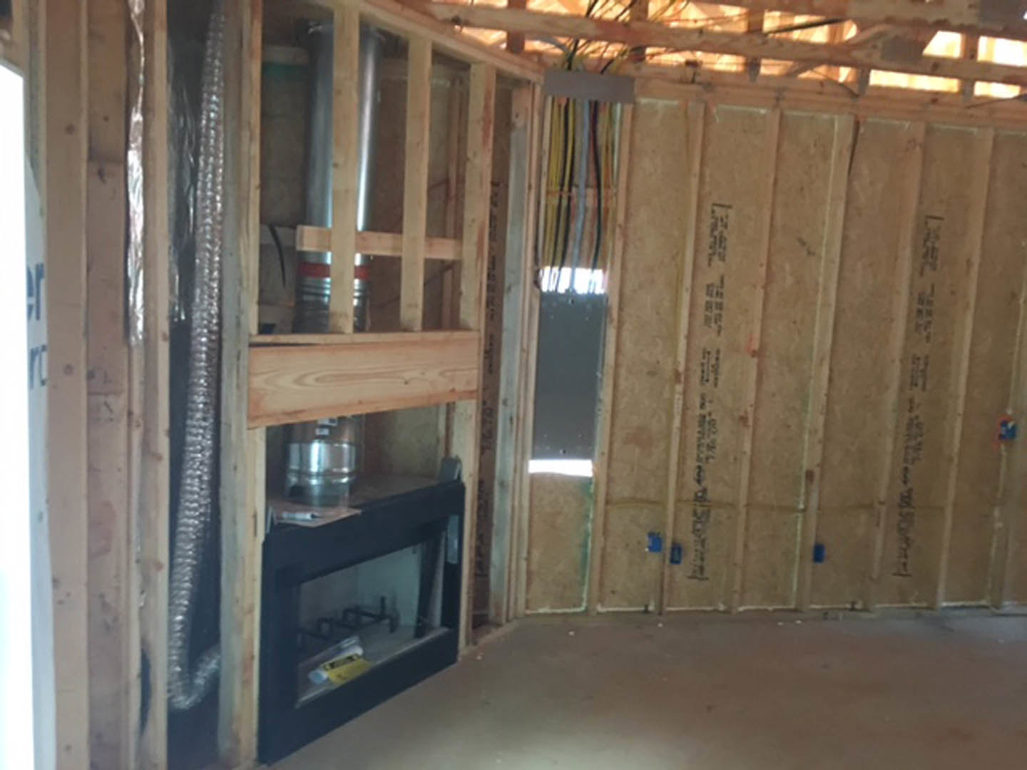 Wood-framed room under construction featuring a black glass-door fireplace, exposed beams with wiring, unfinished walls, and construction materials scattered on the floor
