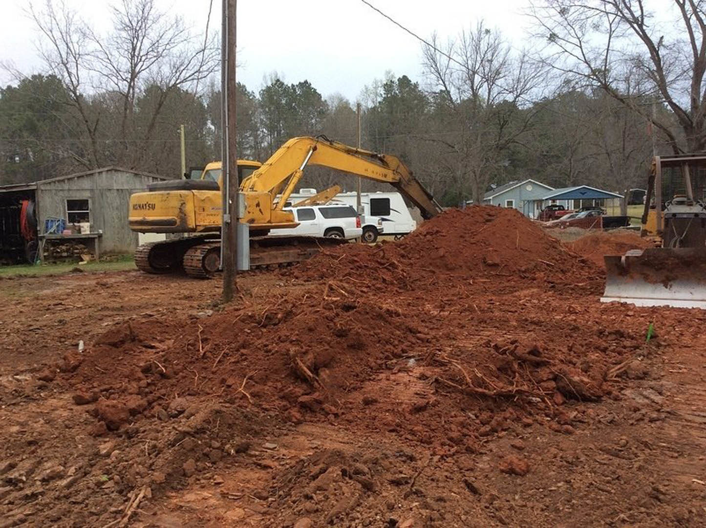 Yellow excavator atop a large dirt pile in front of a partially constructed house with white-framed windows, surrounded by soil and scattered construction materials.