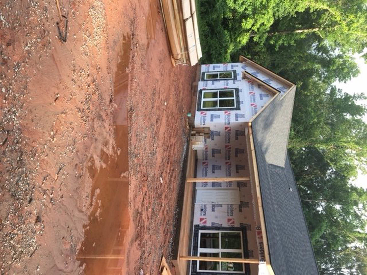 Two-story house under construction with exposed wooden framing, large windows, muddy ground, and surrounding trees