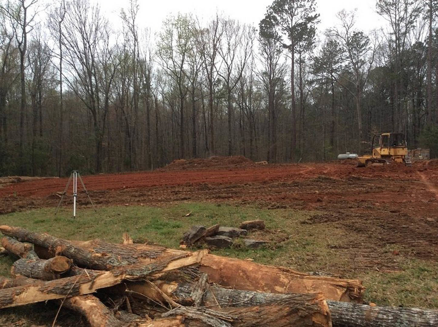 Construction site with a yellow tractor on soil, pile of logs on grassy area, leafless trees in background under blue sky