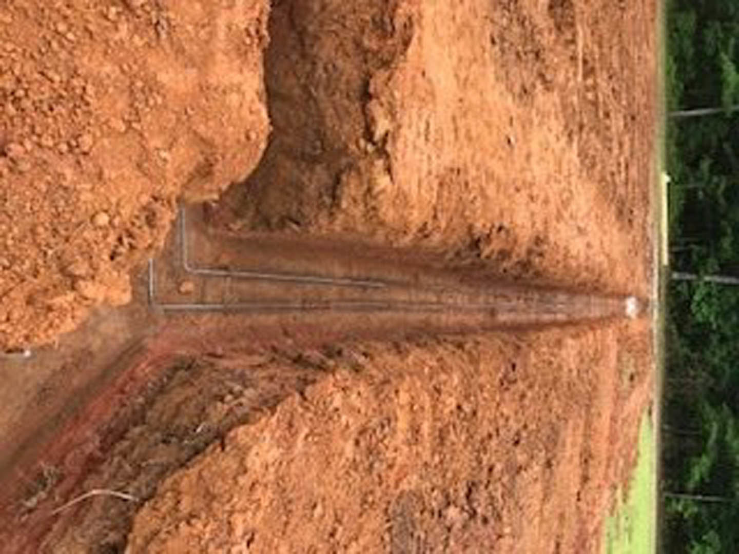 Long stretch of exposed soil with scattered rocks and pipes, bordered by grass and plants in an outdoor residential setting
