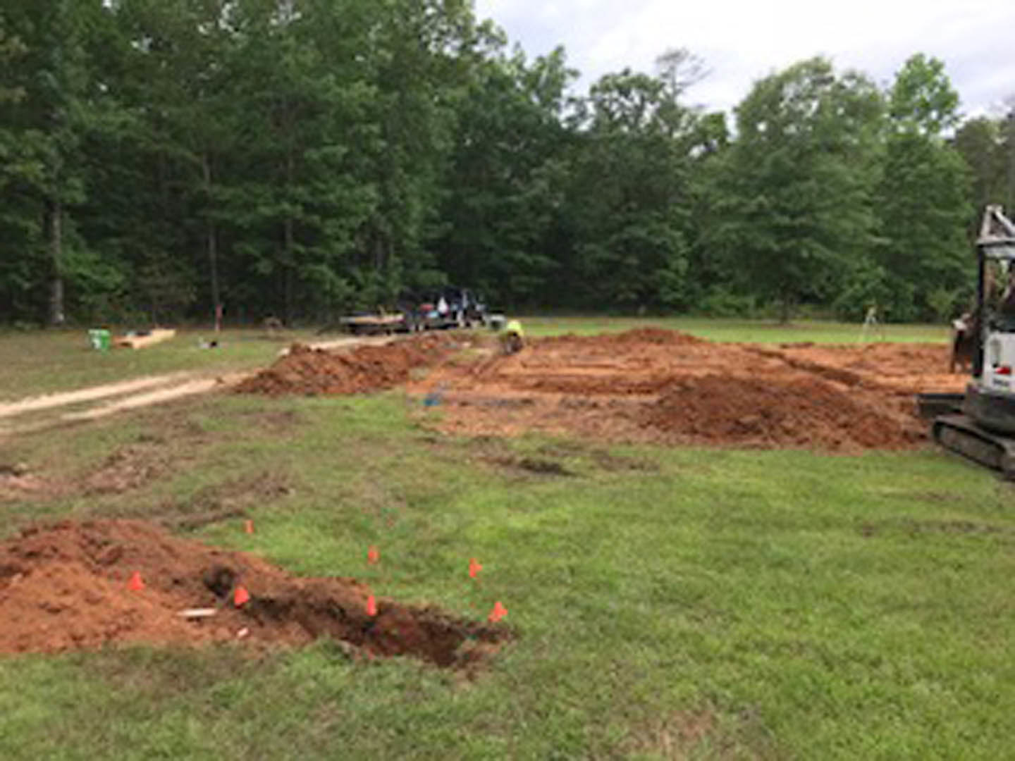 Dirt construction site bordered by mature trees, orange cones marking boundaries, grassy patches visible, overcast sky above