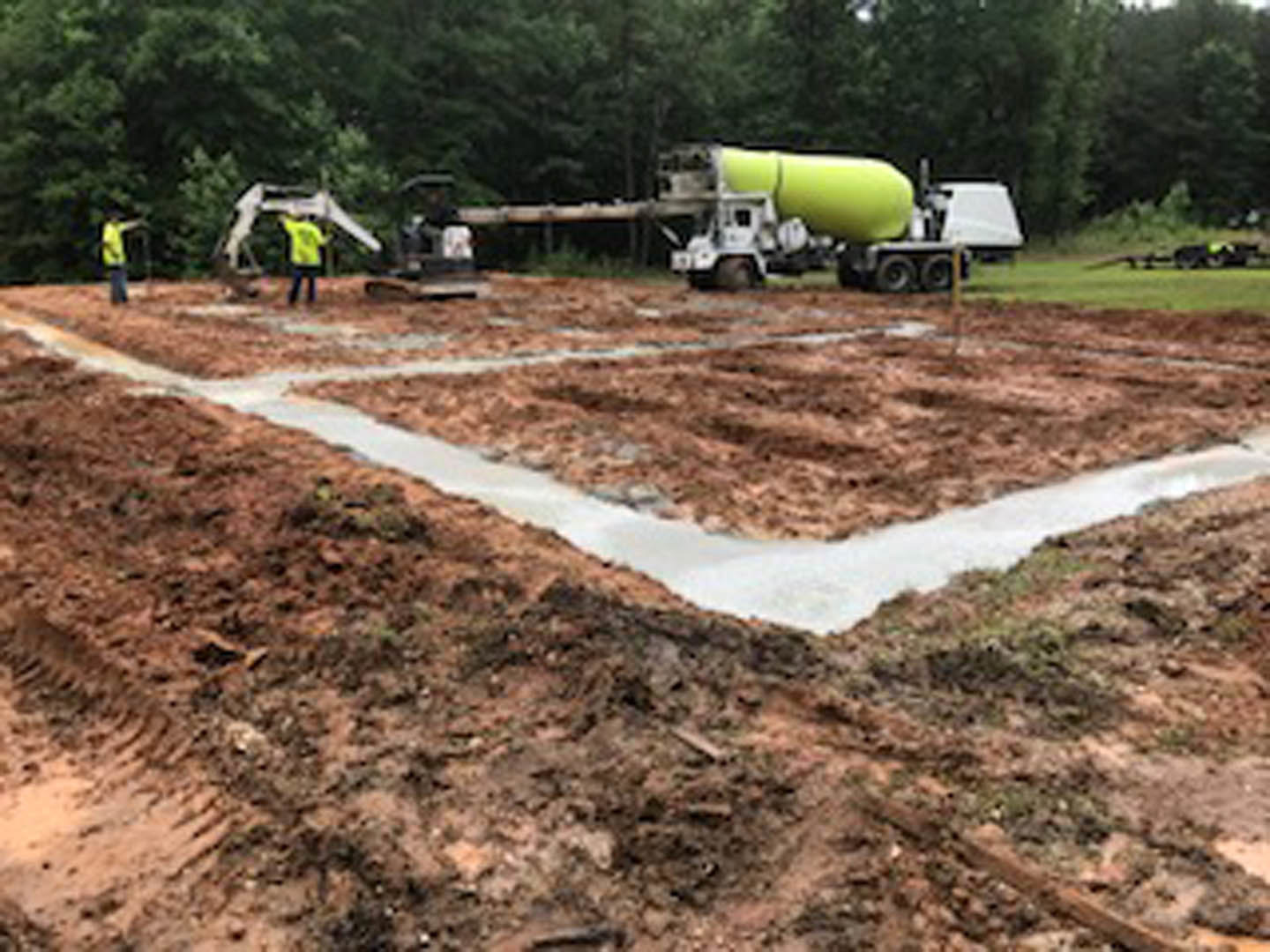 Cement mixer truck and pickup truck parked on dirt construction site, surrounded by grass, trees, and construction materials; worker in yellow shirt near large yellow tank and