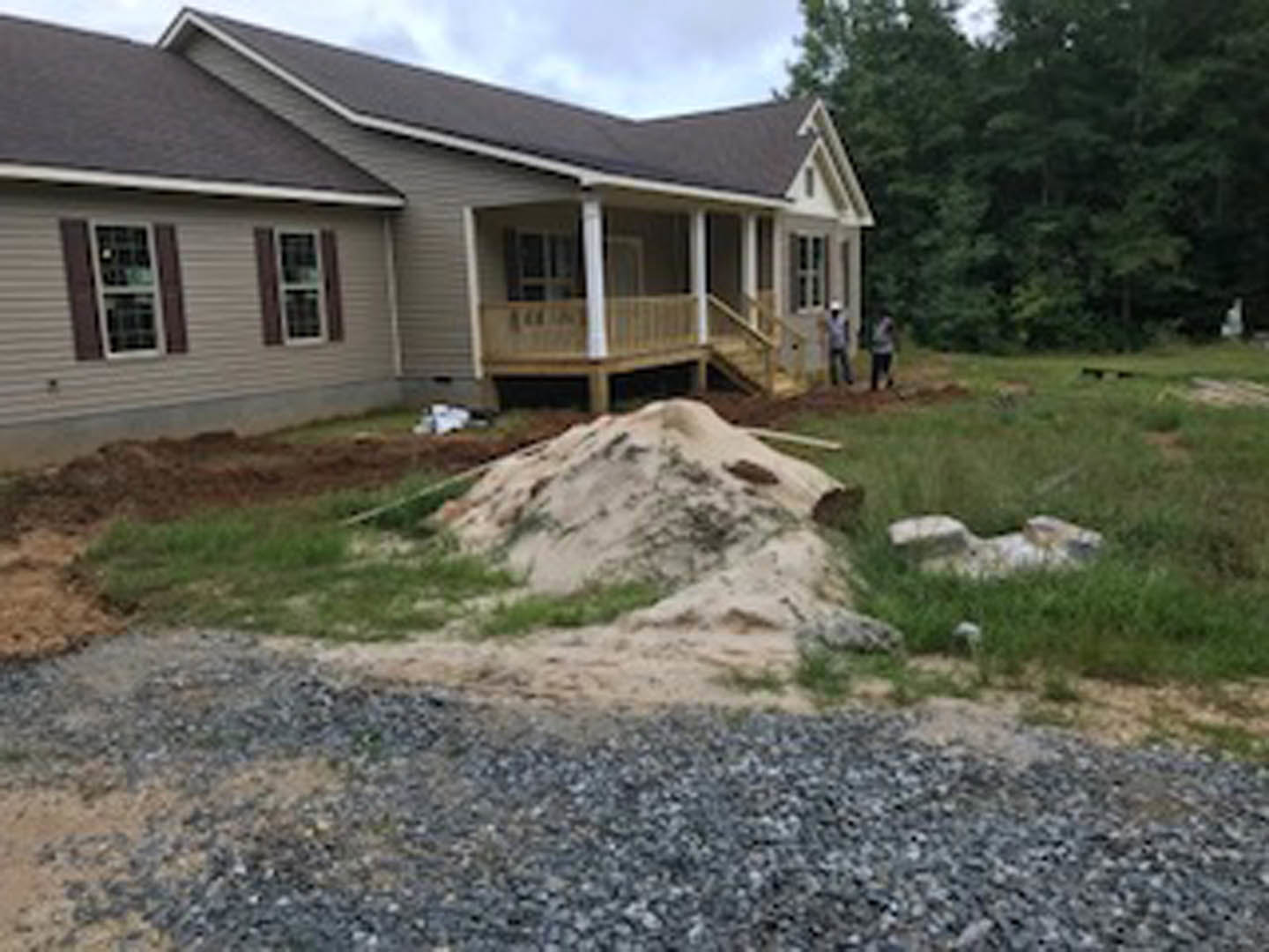 Two-story cottage-style home with tall roof, surrounded by trees, piles of dirt and grass in front yard, gravel driveway, wooden railing, window displaying a sign