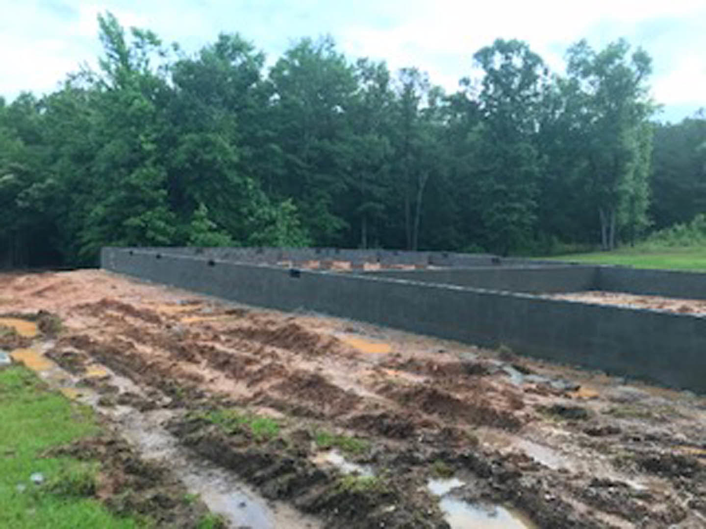 Concrete house foundation surrounded by dirt and grass, bordered by trees and a partially visible road with a concrete wall under a cloudy sky