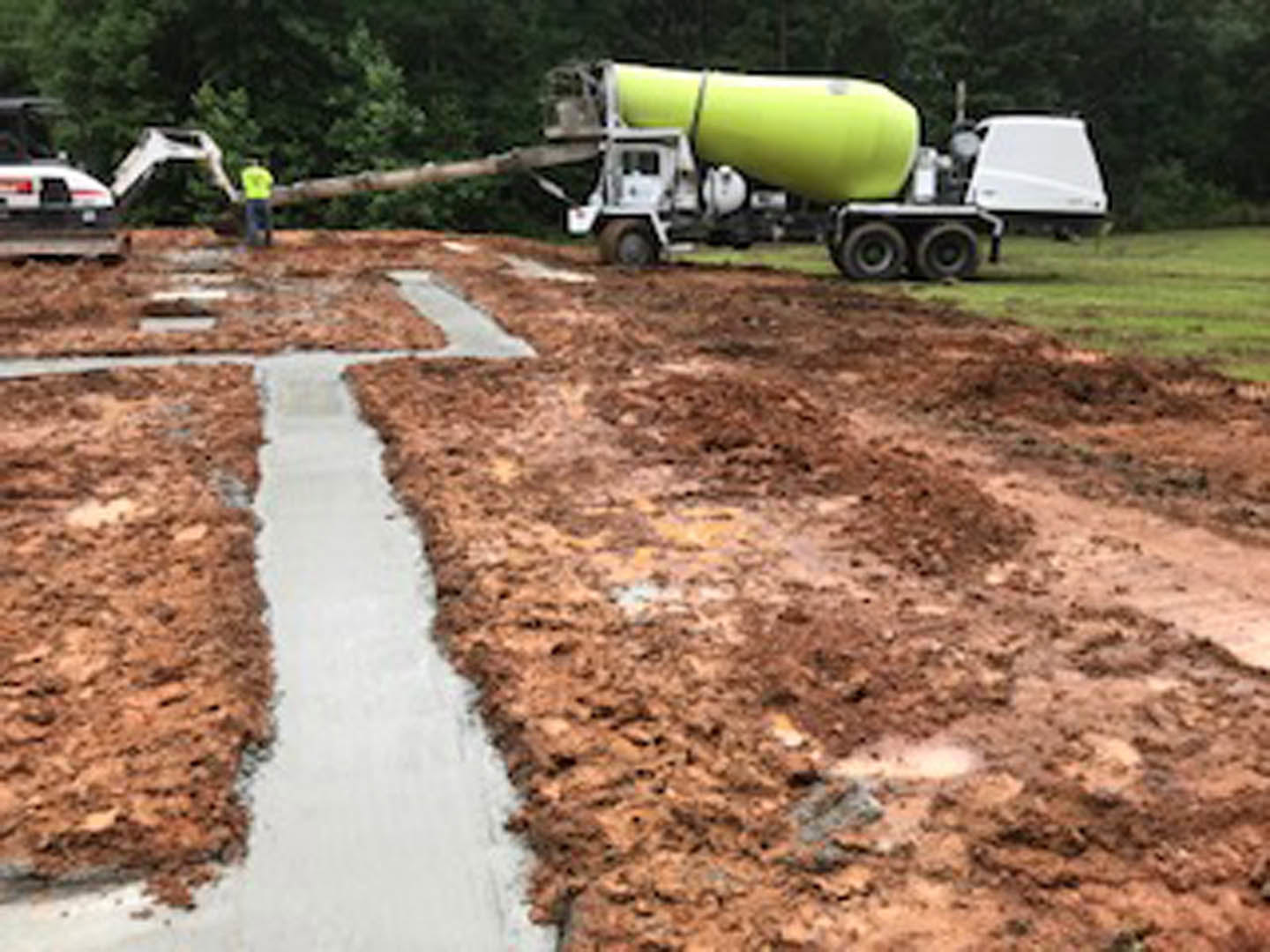 Concrete mixer truck parked on dirt lot surrounded by grass and trees, large cylindrical drum visible, construction materials and soil in foreground