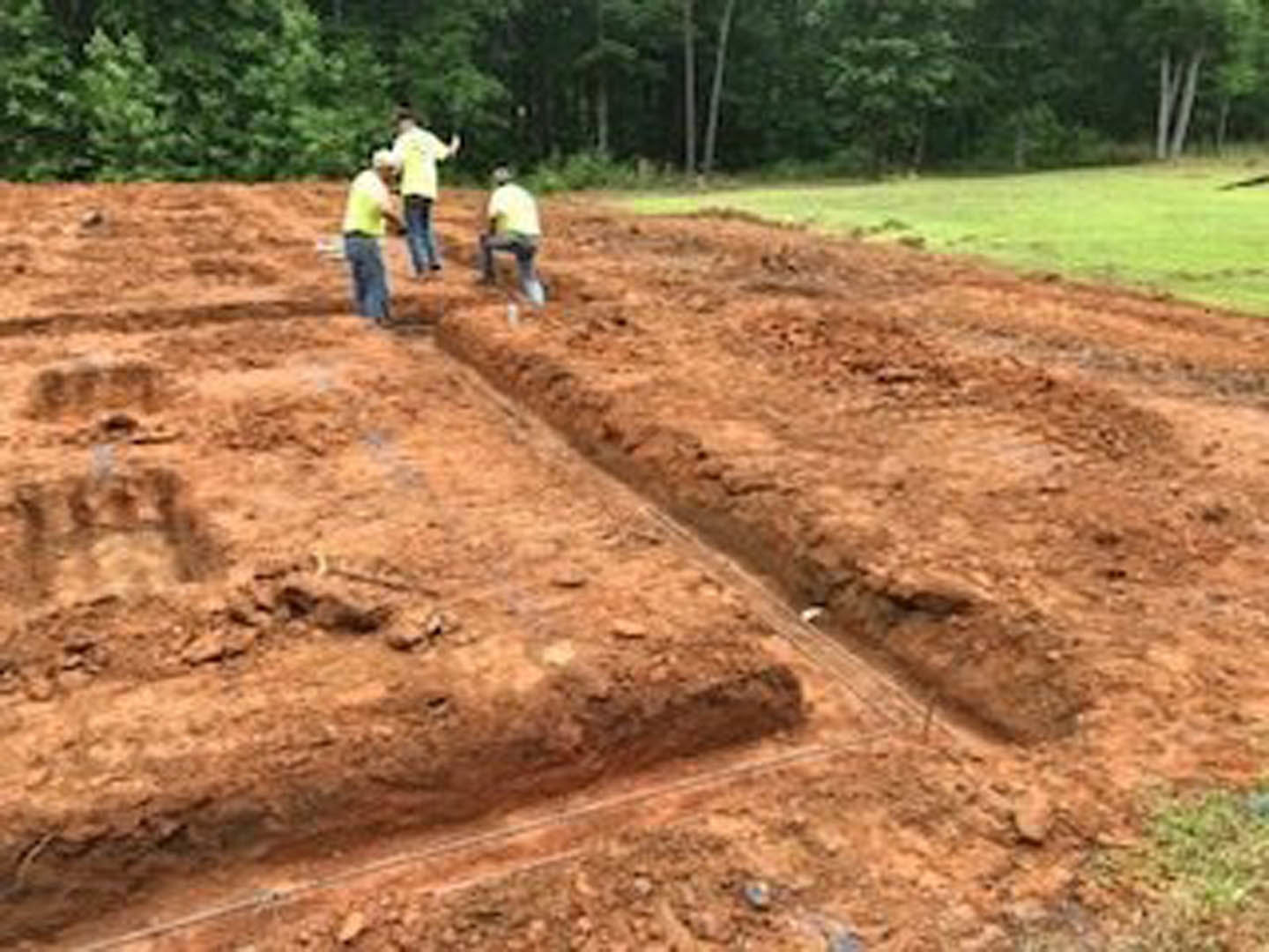 Construction crew preparing muddy foundation in open dirt field bordered by trees and grass