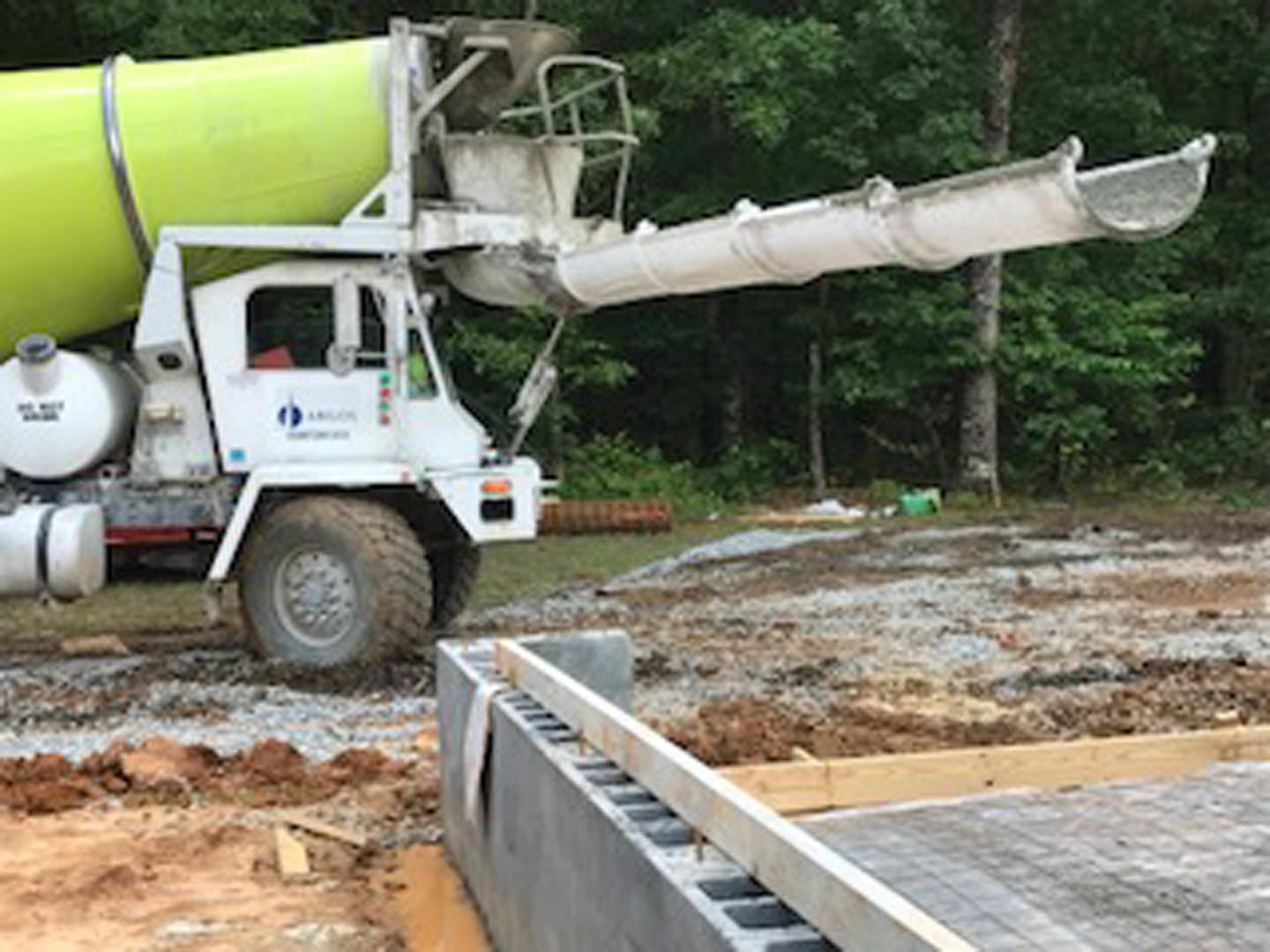 White cement mixer truck parked on dirt near concrete slab with wooden beam, large green and yellow tubes attached, trees in background, partial logo visible on container.