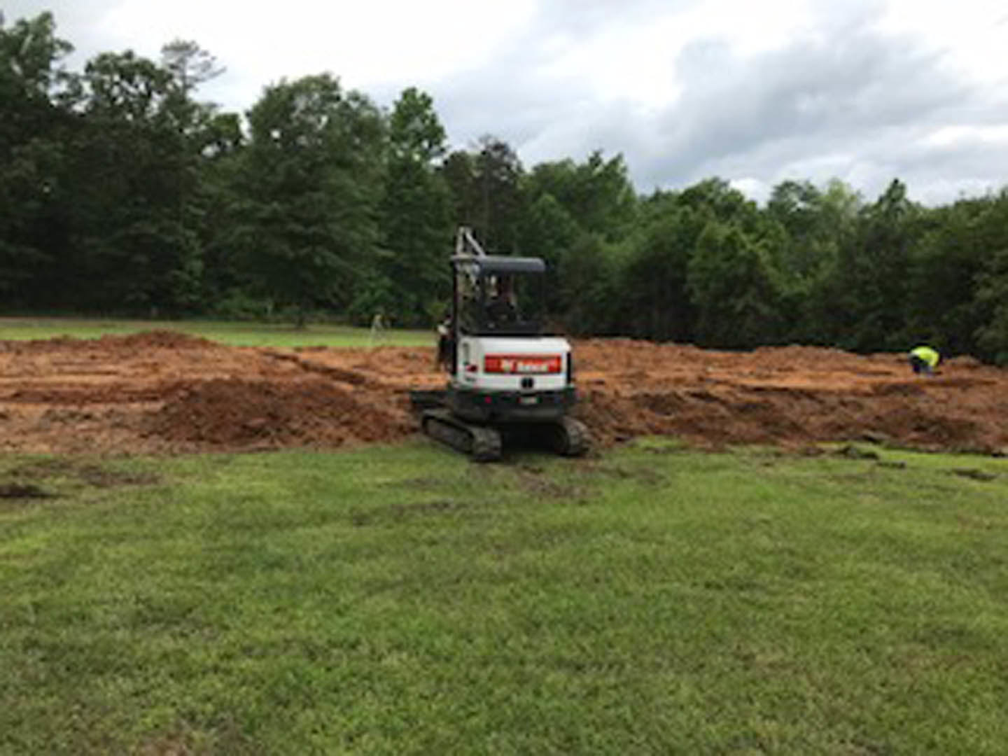 Yellow bulldozer parked on grassy field with distant trees under blue sky, soil exposed in foreground