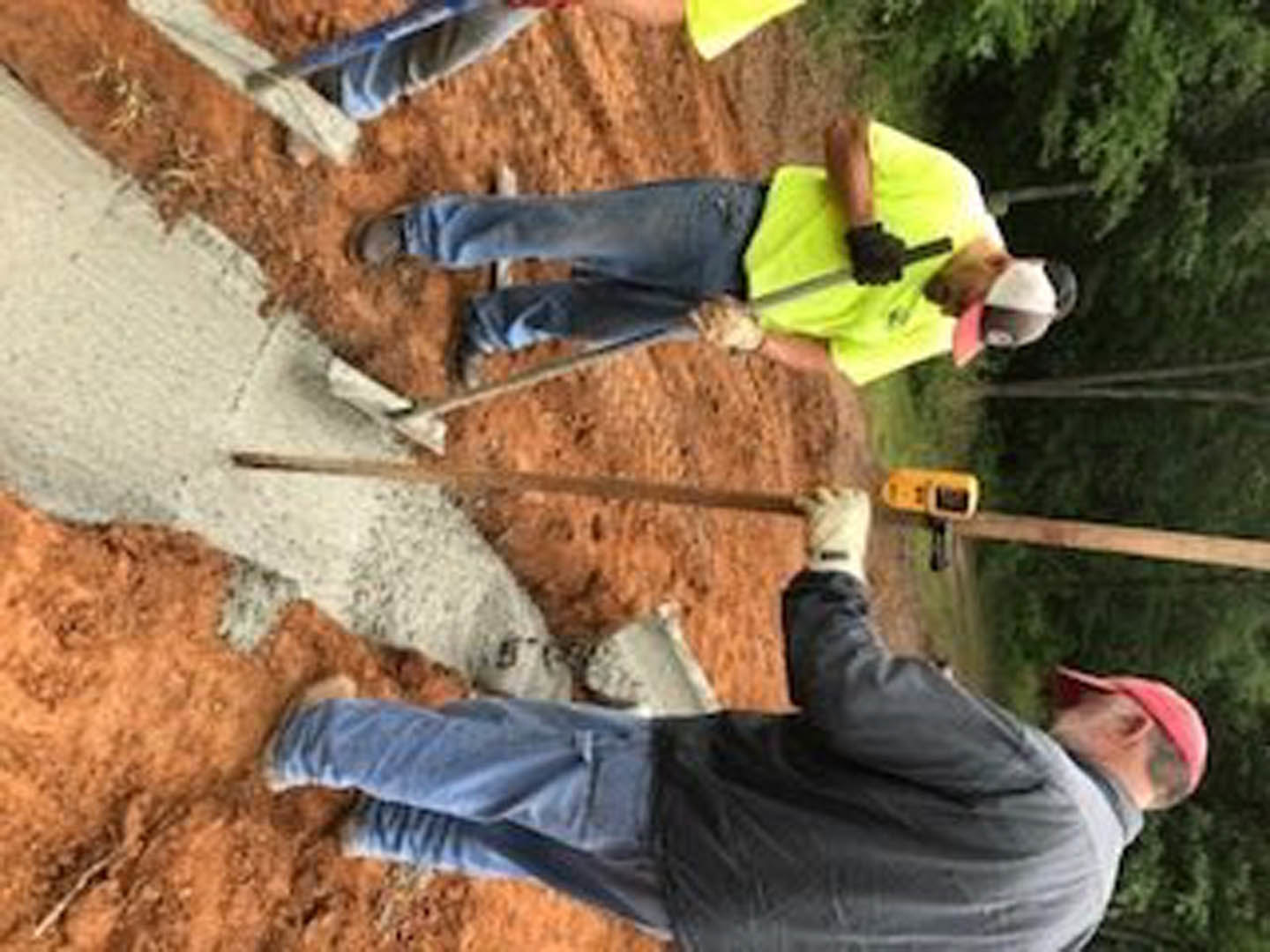 Men in high-visibility clothing and hard hats leveling concrete on a sidewalk outside a custom home construction site, surrounded by trees and dirt ground.