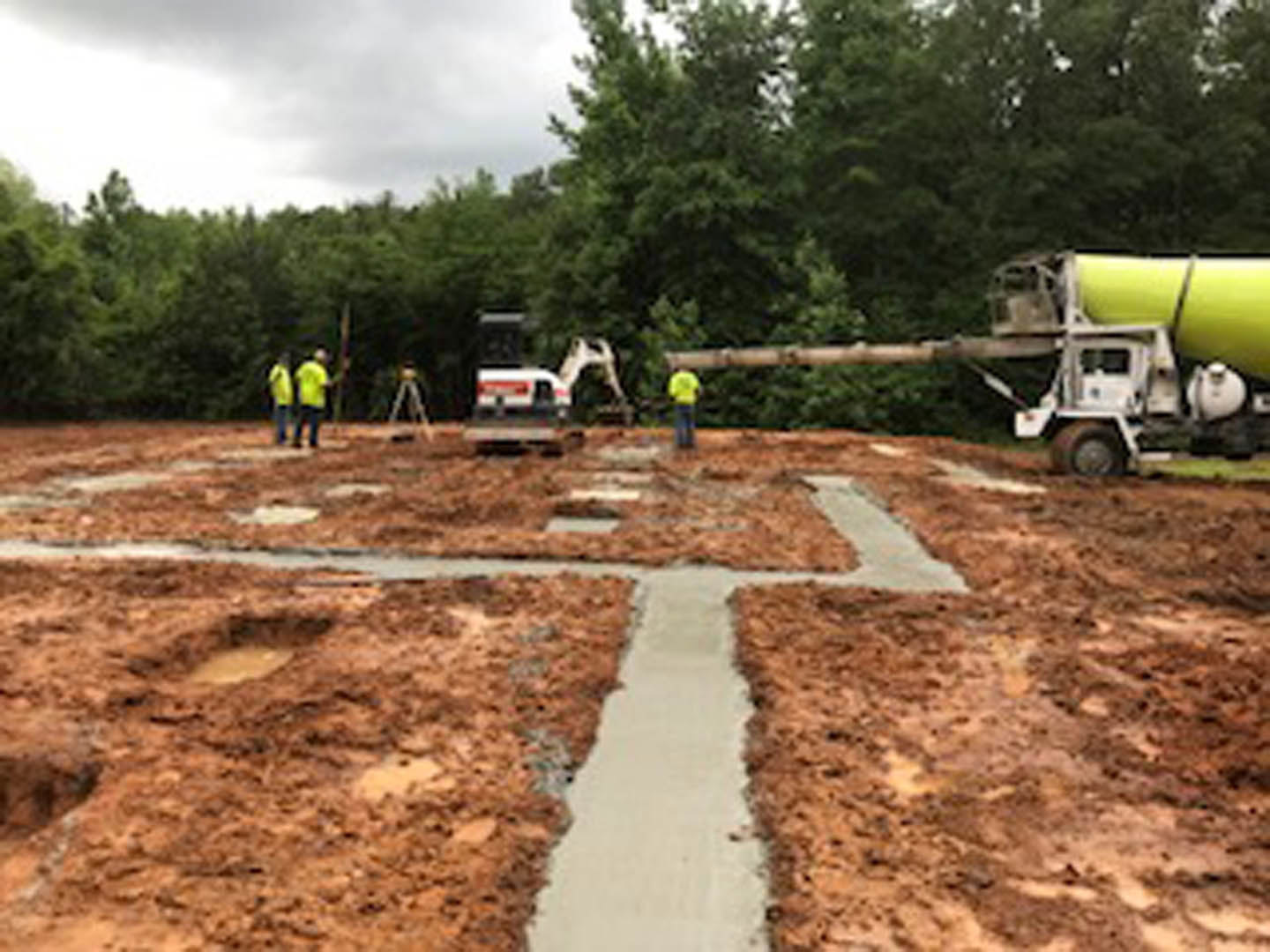 Men mixing concrete beside a white and red truck with a large yellow cylinder, concrete path extending through grassy field, trees in background