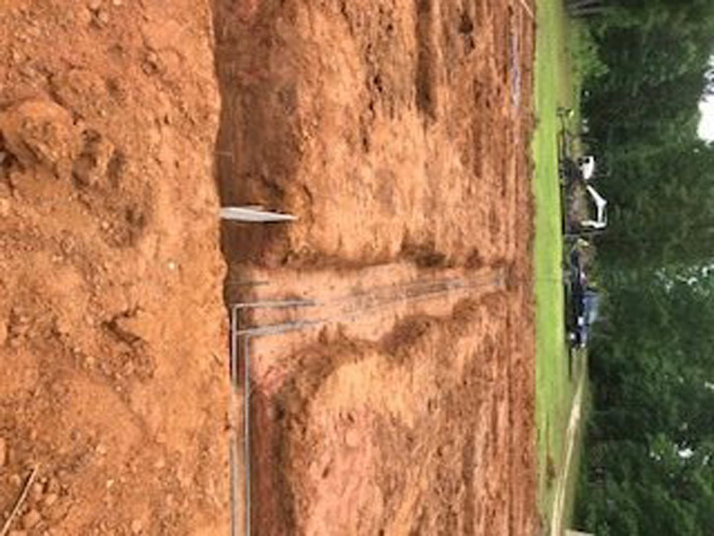 Dirt field with scattered metal pipes, grassy area, rock wall in foreground, road and trees in background