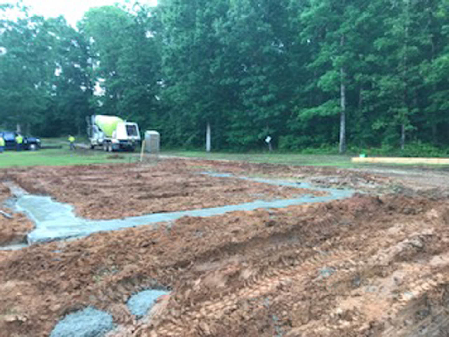 Cement mixer truck parked on dirt construction site surrounded by green trees, muddy ground, and grassy field; worker in yellow vest visible in background.