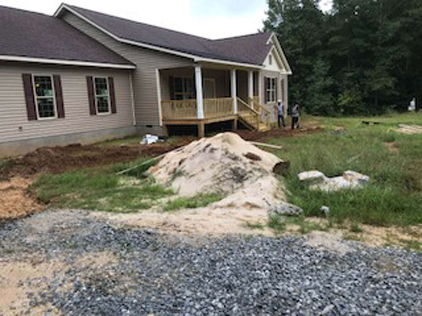 Two-story house with white-framed windows, surrounded by trees and grass, pile of dirt and gravel in the yard, person sitting on a bench in the background