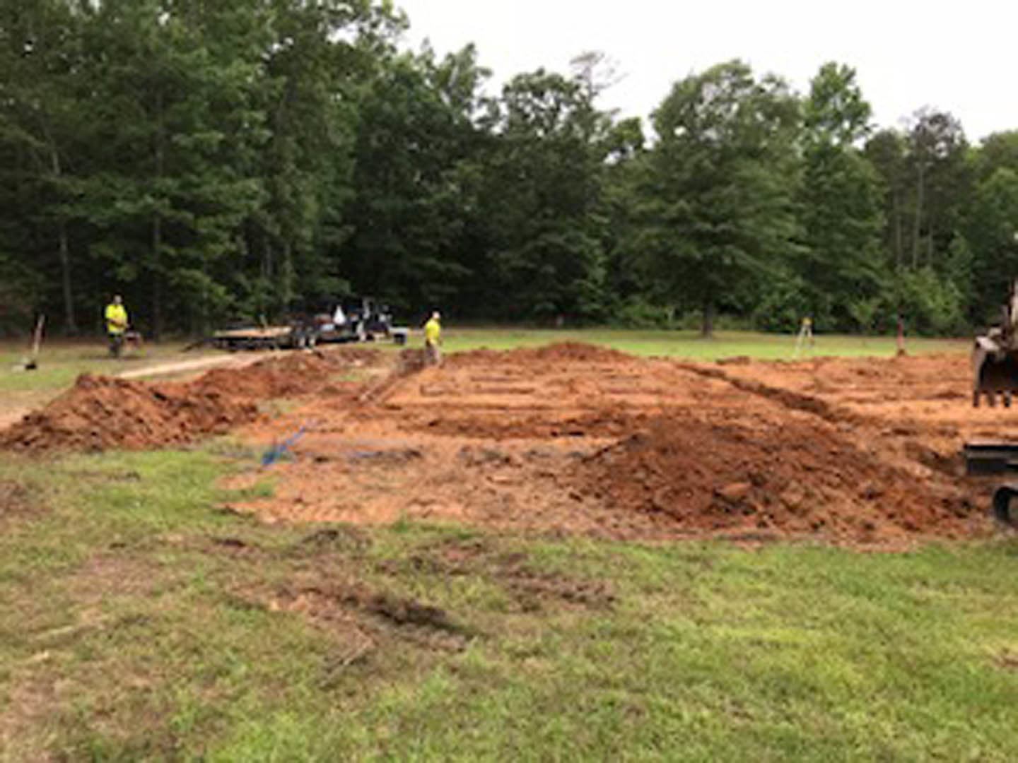 Partially built custom home surrounded by trees, exposed soil and grass, construction workers standing on muddy ground