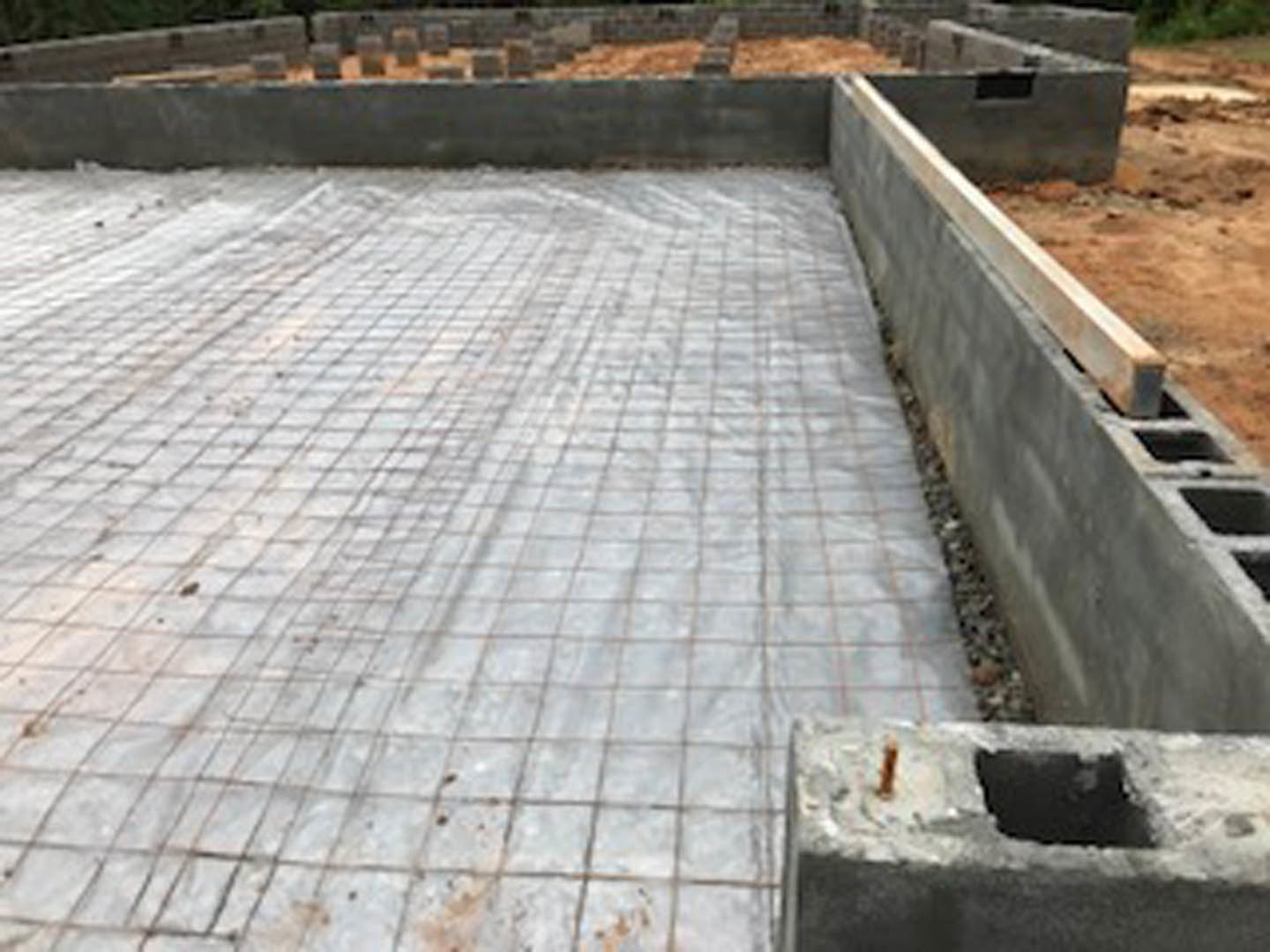Concrete foundation with stacked cement blocks and exposed wooden beam, surrounded by construction materials on outdoor ground.