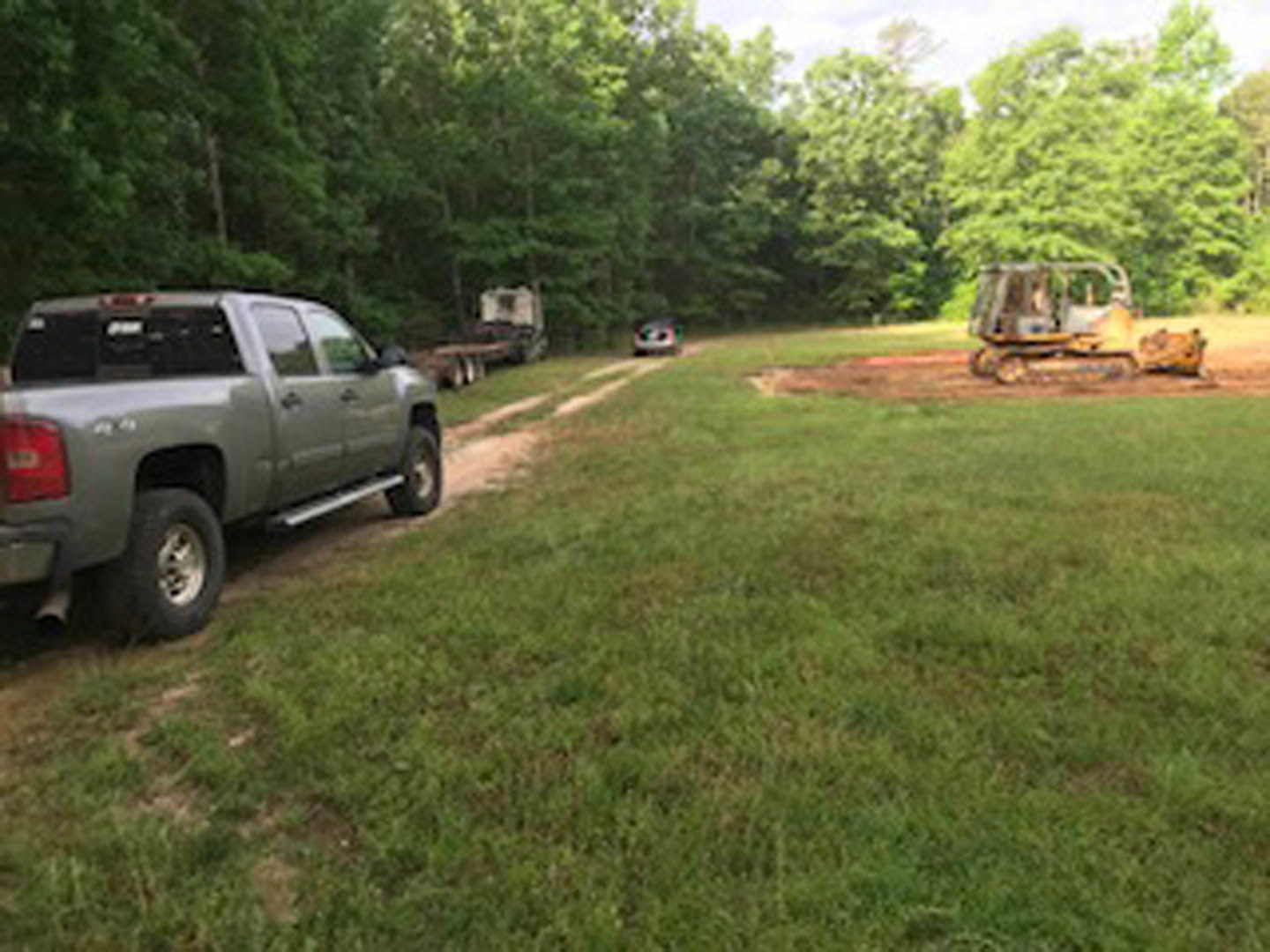 White pickup truck parked on grassy lawn near dense trees, dirt road visible, blurred yellow tractor and trailer in background