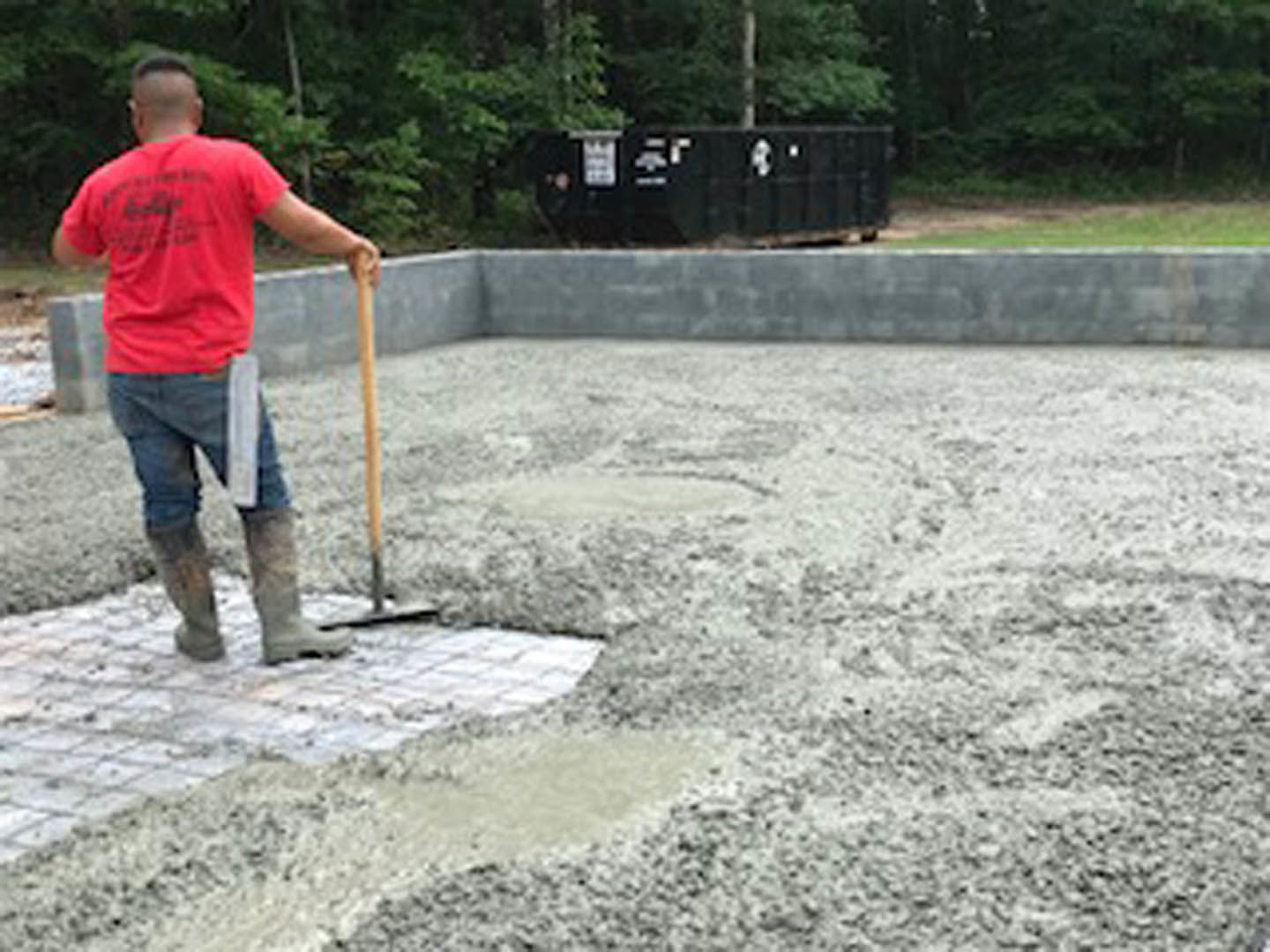 Man in red shirt holding shovel and paper, standing on dirt ground near black wall with white signs, surrounded by trees and concrete.