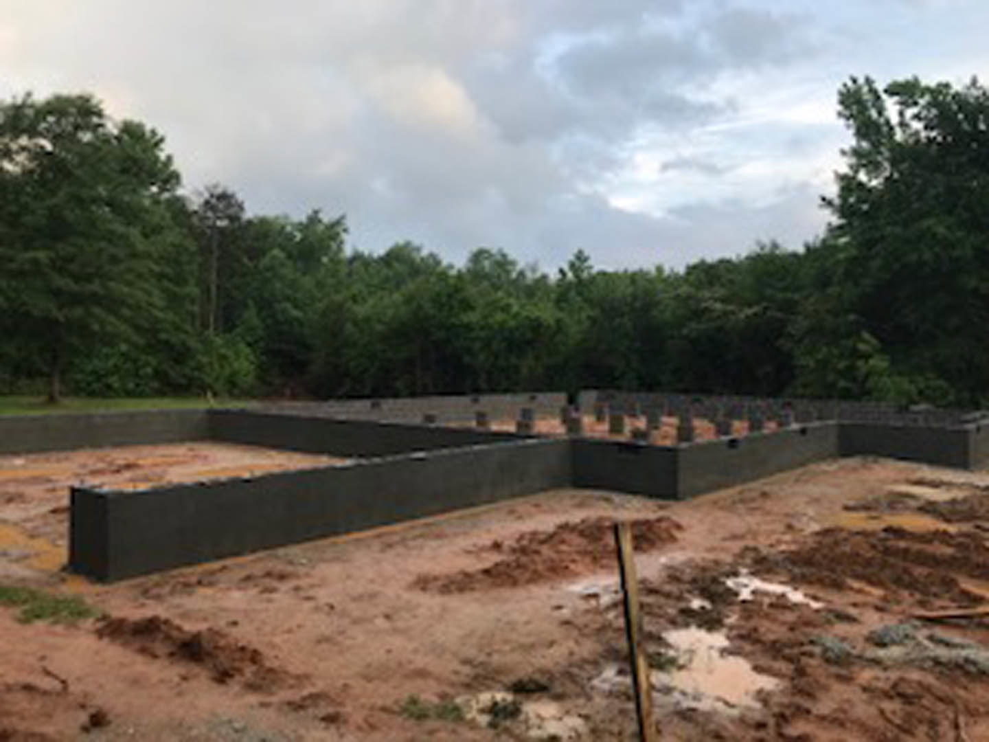 Concrete foundation wall with exposed soil and dirt lot, distant trees and cloudy sky, partial view of fence and utility pole