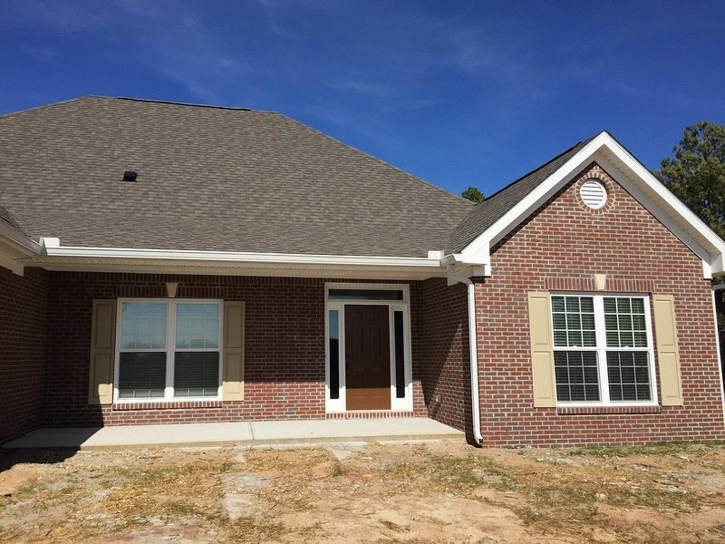 Red brick house with white-trimmed brown door, windows with blinds, landscaped green yard, and visible exterior vent under cloudy sky