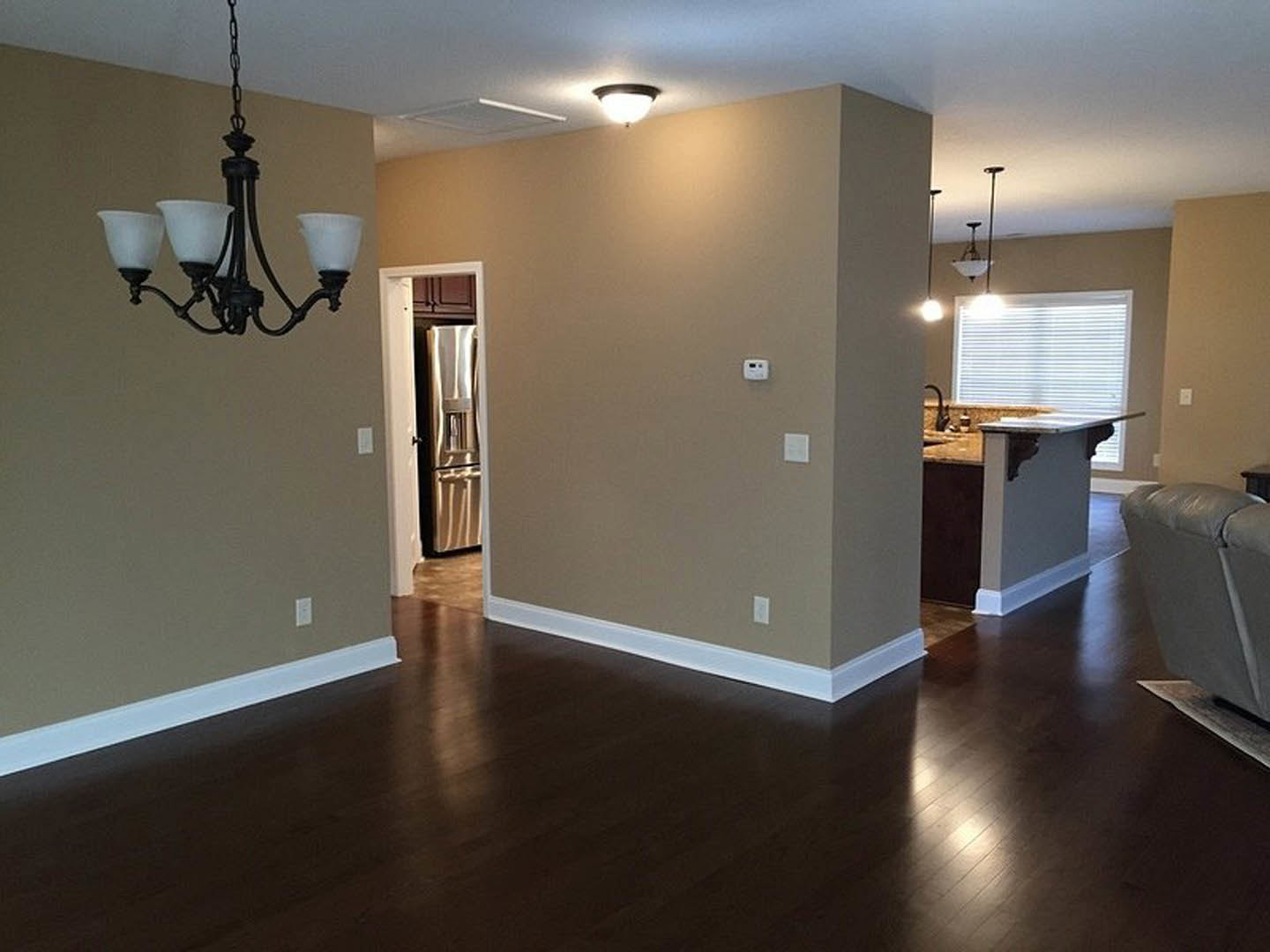 Open-concept living room with wood flooring, white kitchen cabinets, black-edged countertop, modern chandelier, plush couch, and decorative lamp.