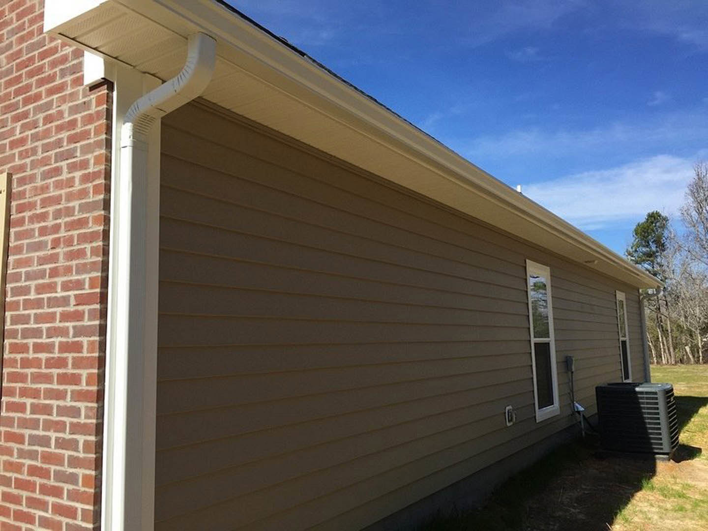Red brick exterior wall with white-framed window, black air conditioner unit on grass, gray shingle roof, leafy tree nearby under partly cloudy sky