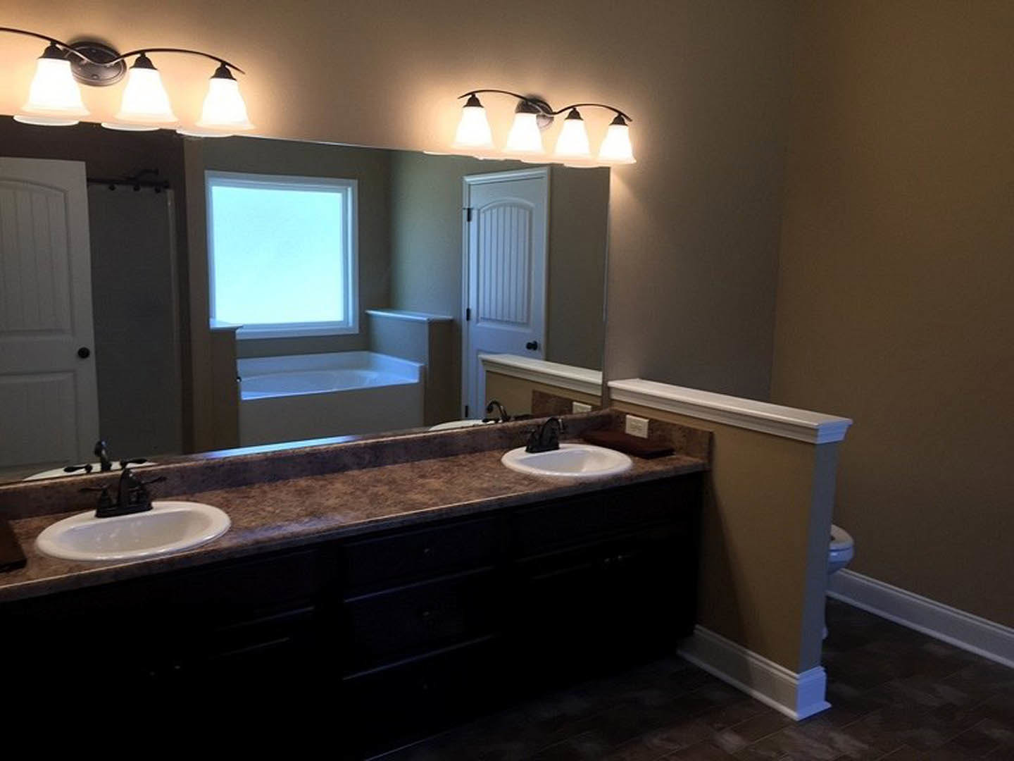 Bathroom with expansive mirror above white countertop and modern sink, wall-mounted light fixture, black soap dispenser, tiled backsplash, and window with natural light.