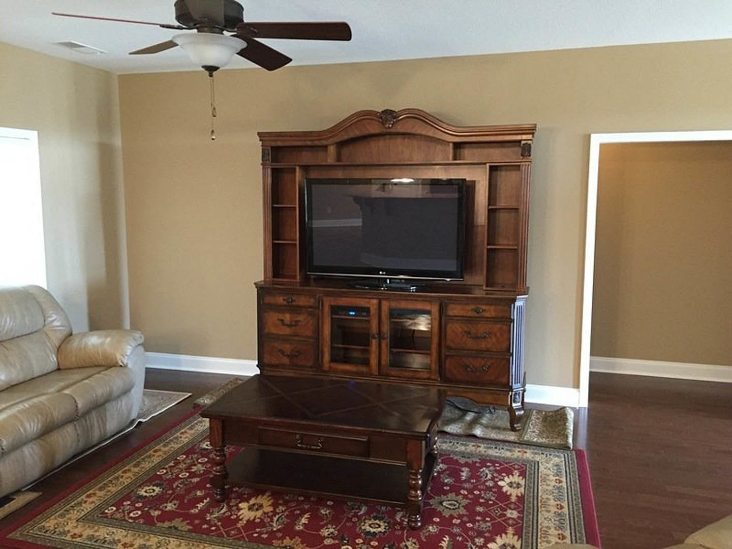 Living room with wood flooring, large wall-mounted TV above a stone fireplace, ceiling fan, coffee table, and neutral-toned couch.