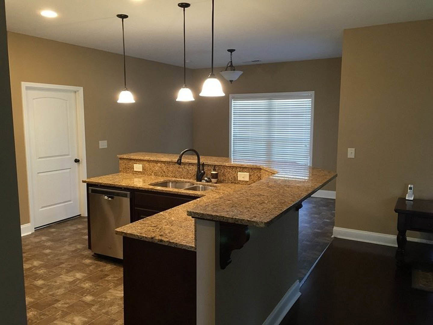 Granite kitchen countertop with undermount sink, tile backsplash, white cabinetry, pendant lights, window with blinds, and white door with black handle