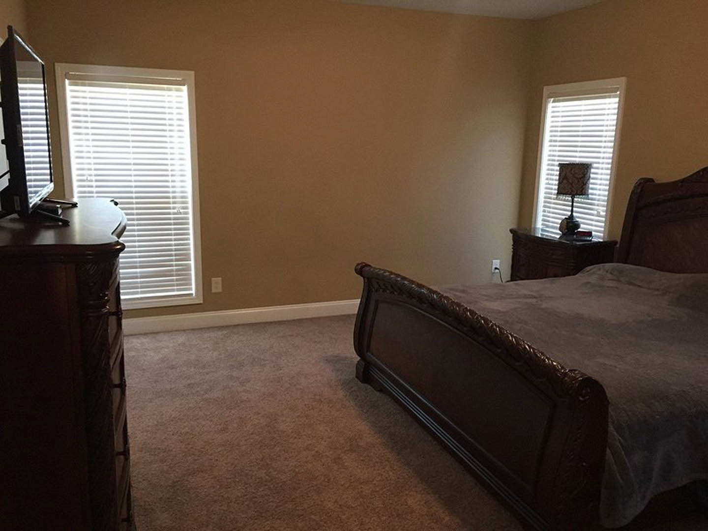 Bedroom with upholstered bed, white bedding, wooden dresser, desk positioned in front of large window with sheer curtains, table lamp with fabric shade, hardwood flooring
