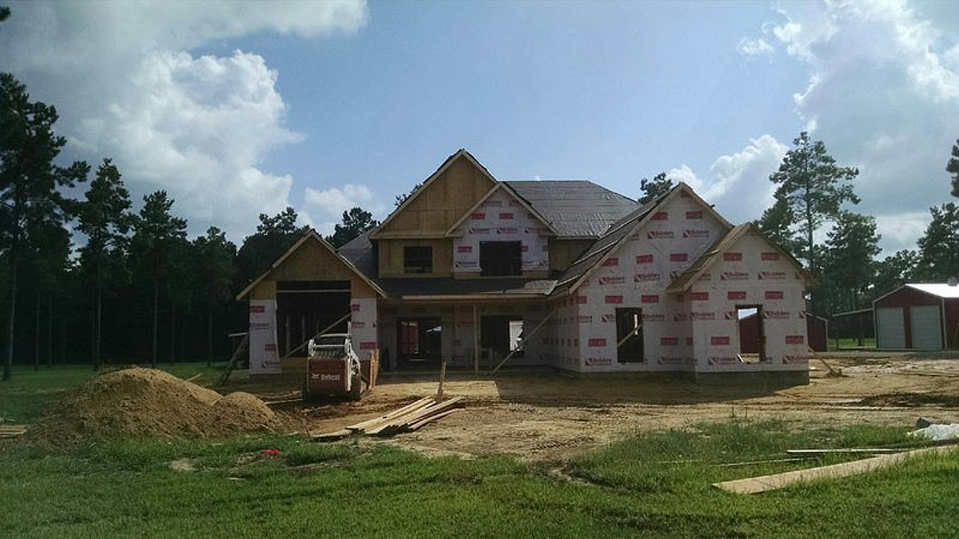 Partially built house with exposed red and white insulation, construction truck parked in front, red shed with white doors nearby, grassy field scattered with dirt and wood, cloudy