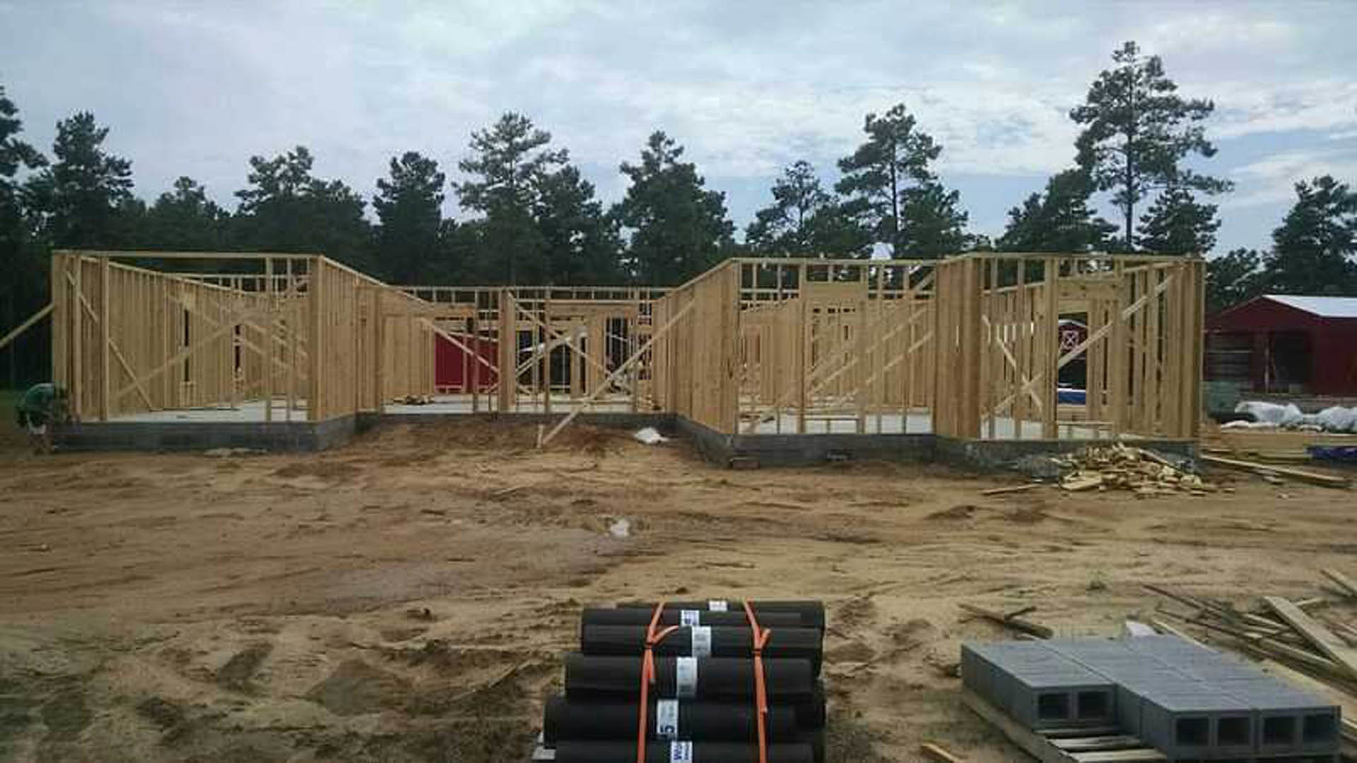 Wood-framed house under construction with scaffolding, stacks of concrete, and black pipes on dirt ground, surrounded by trees in the background