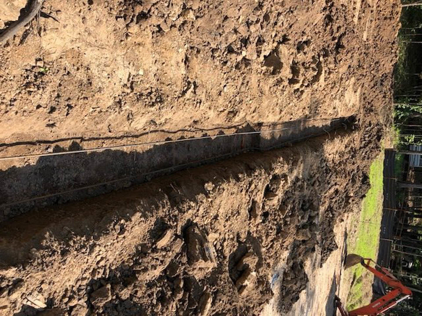 Dirt field with a long metal pipe laid in a trench, red excavator digging nearby, exposed soil and rocks along the trench walls