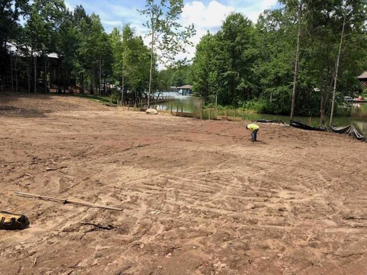 Person in yellow shirt standing on dirt area with leafy trees and a modern house featuring light siding in the background