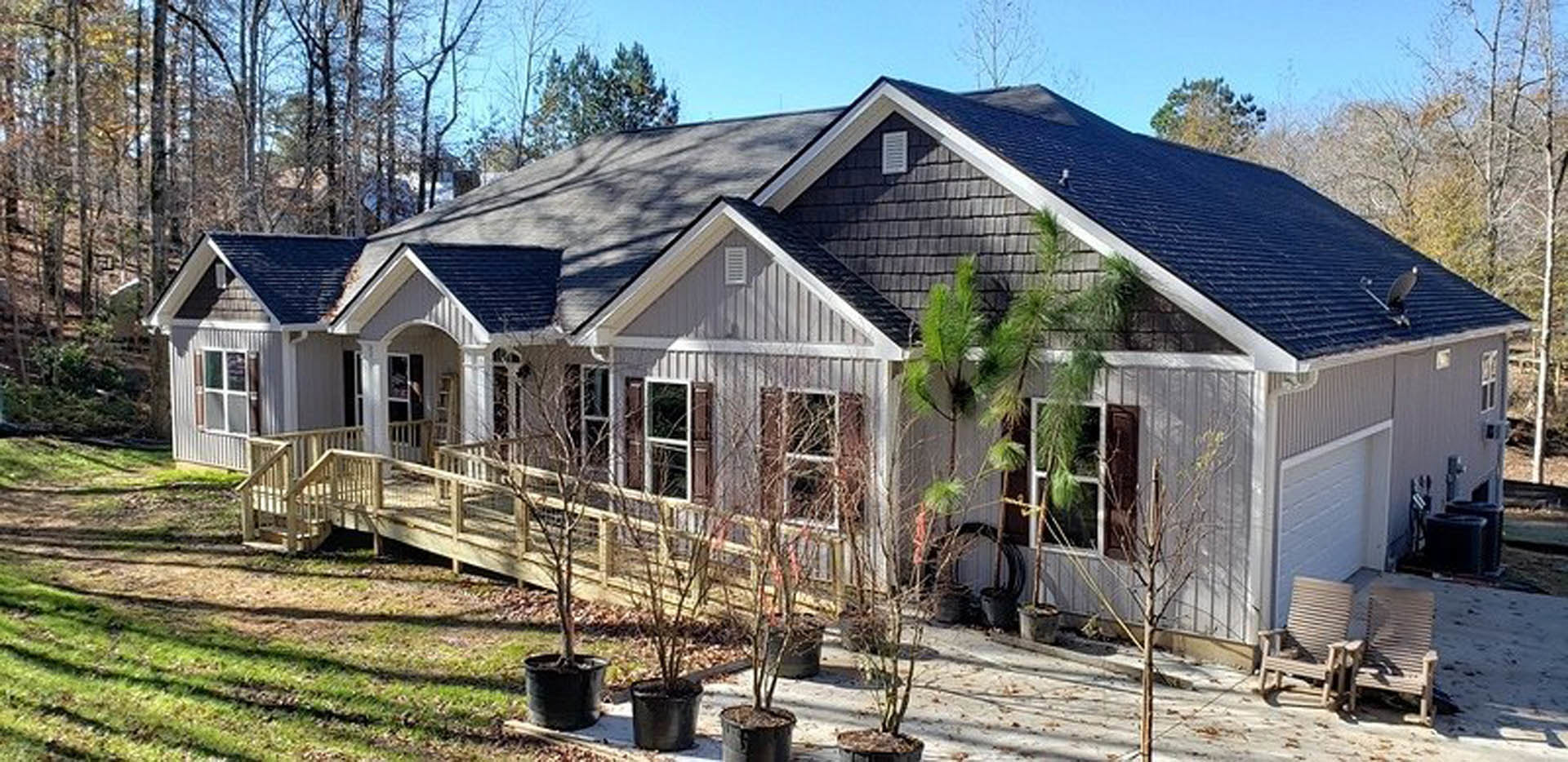 White cottage-style home with covered front porch, black planters, wooden chairs, manicured lawn, and mature trees under a blue sky