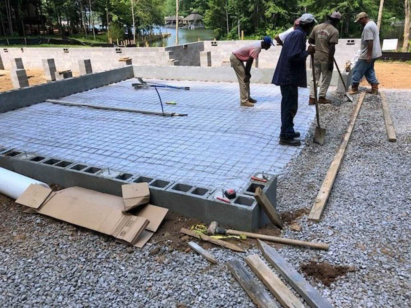 Crew pouring and leveling concrete slab foundation outdoors, surrounded by trees, with workers in various clothing and footwear