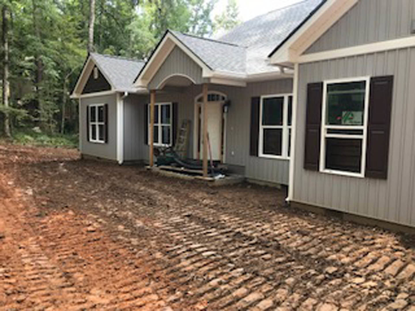 Two-story cottage-style home with light siding, dirt driveway and path, covered porch, white-framed windows, white ladder with sign near entrance, small tree in yard, white cross