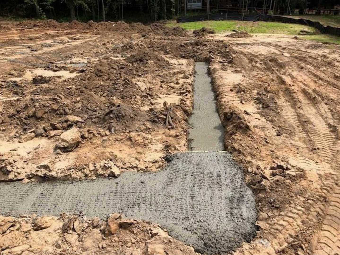 Dirt field with a freshly dug trench, surrounded by scattered rocks and tire tracks, with blurred trees in the background