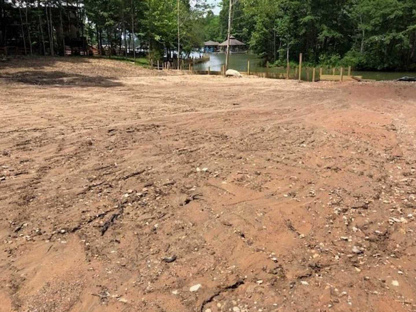 Dirt clearing bordered by mature trees, small body of water visible beyond, natural landscaping with soil and sparse vegetation