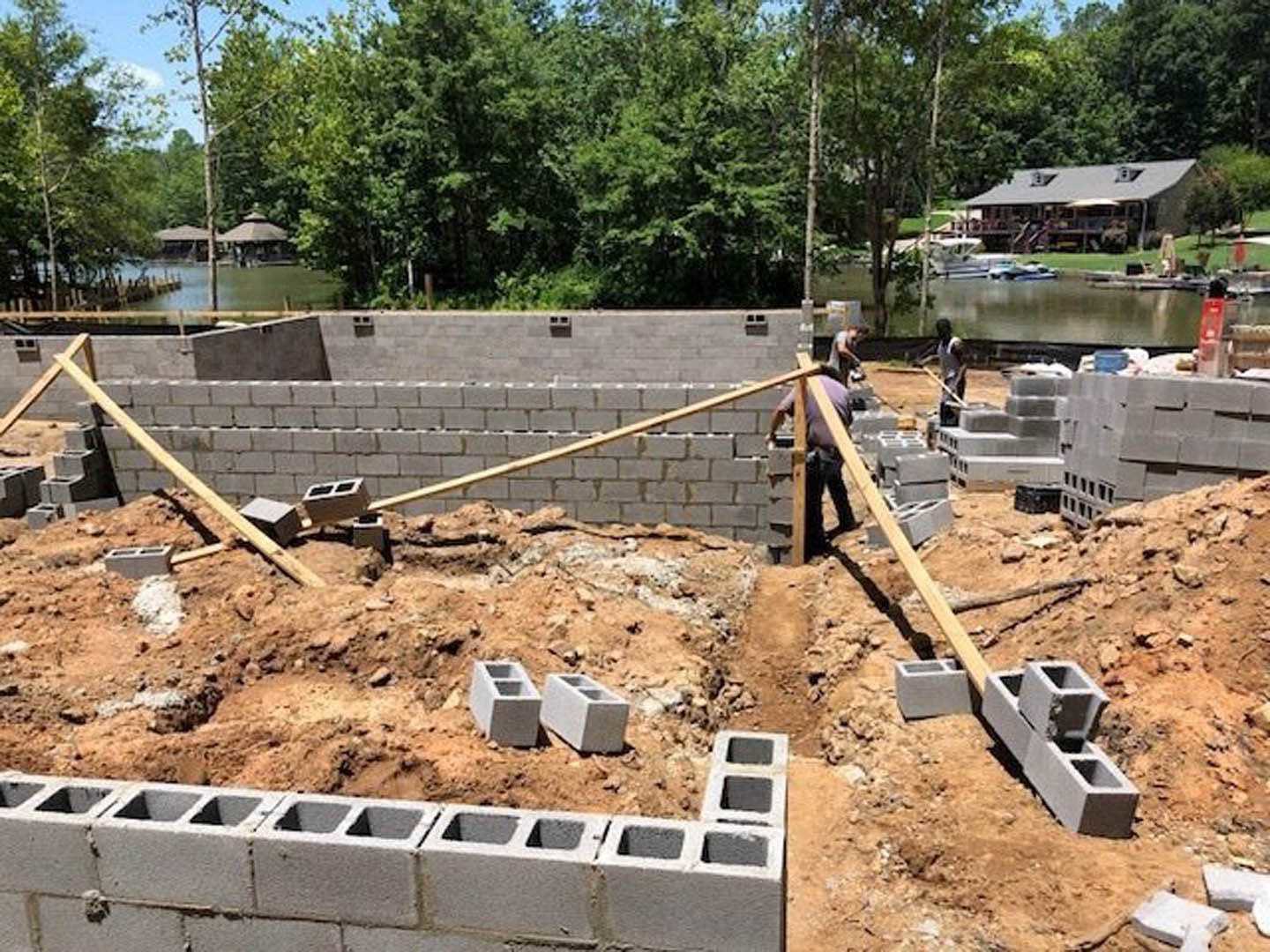 Two workers on a residential construction site with exposed foundation, soil, scattered wooden beams, and a parked car in the background.