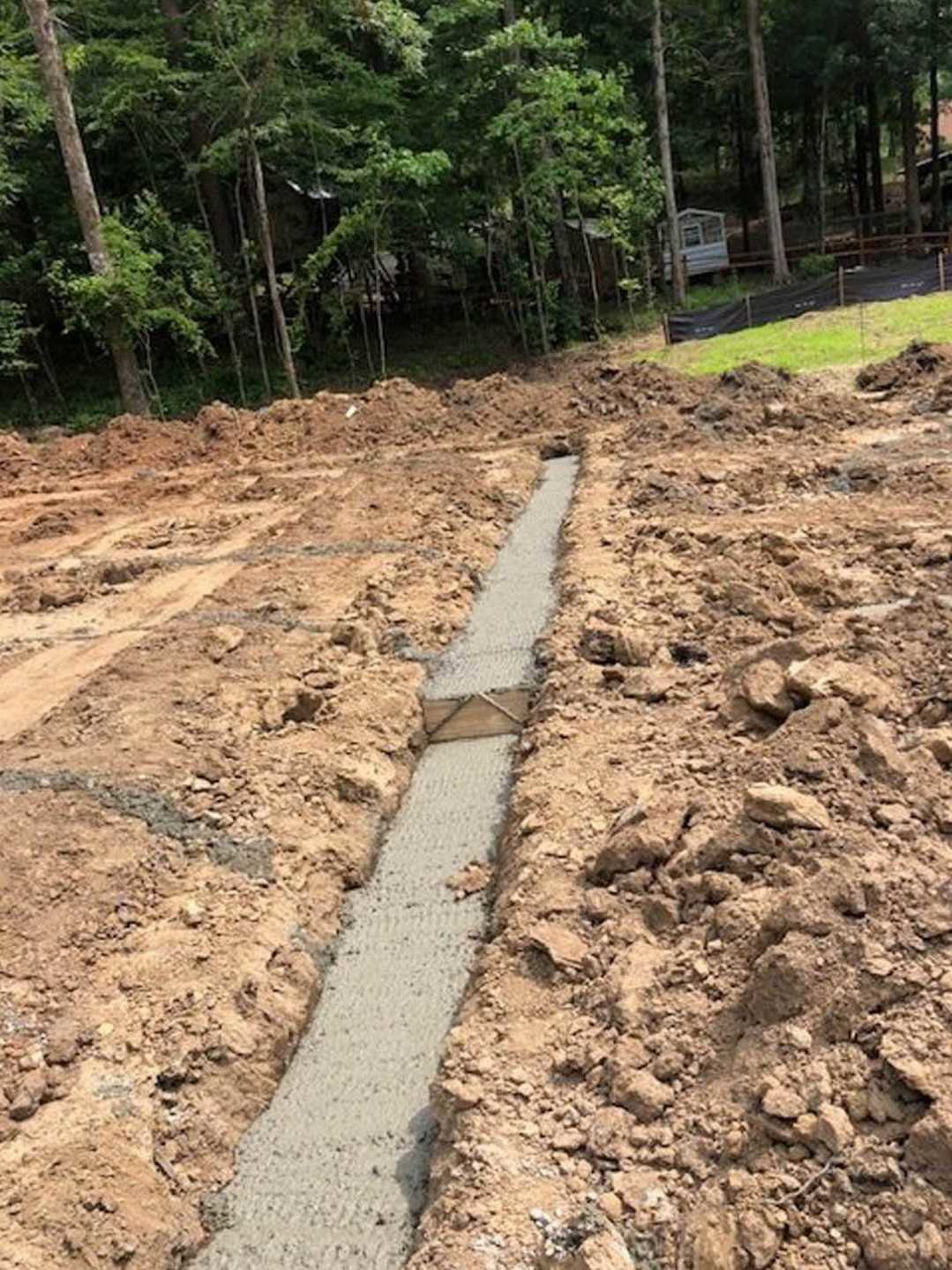 Wide dirt trench bordered by grass and trees, with a wooden fence in the background and a partially visible house nestled among woods.