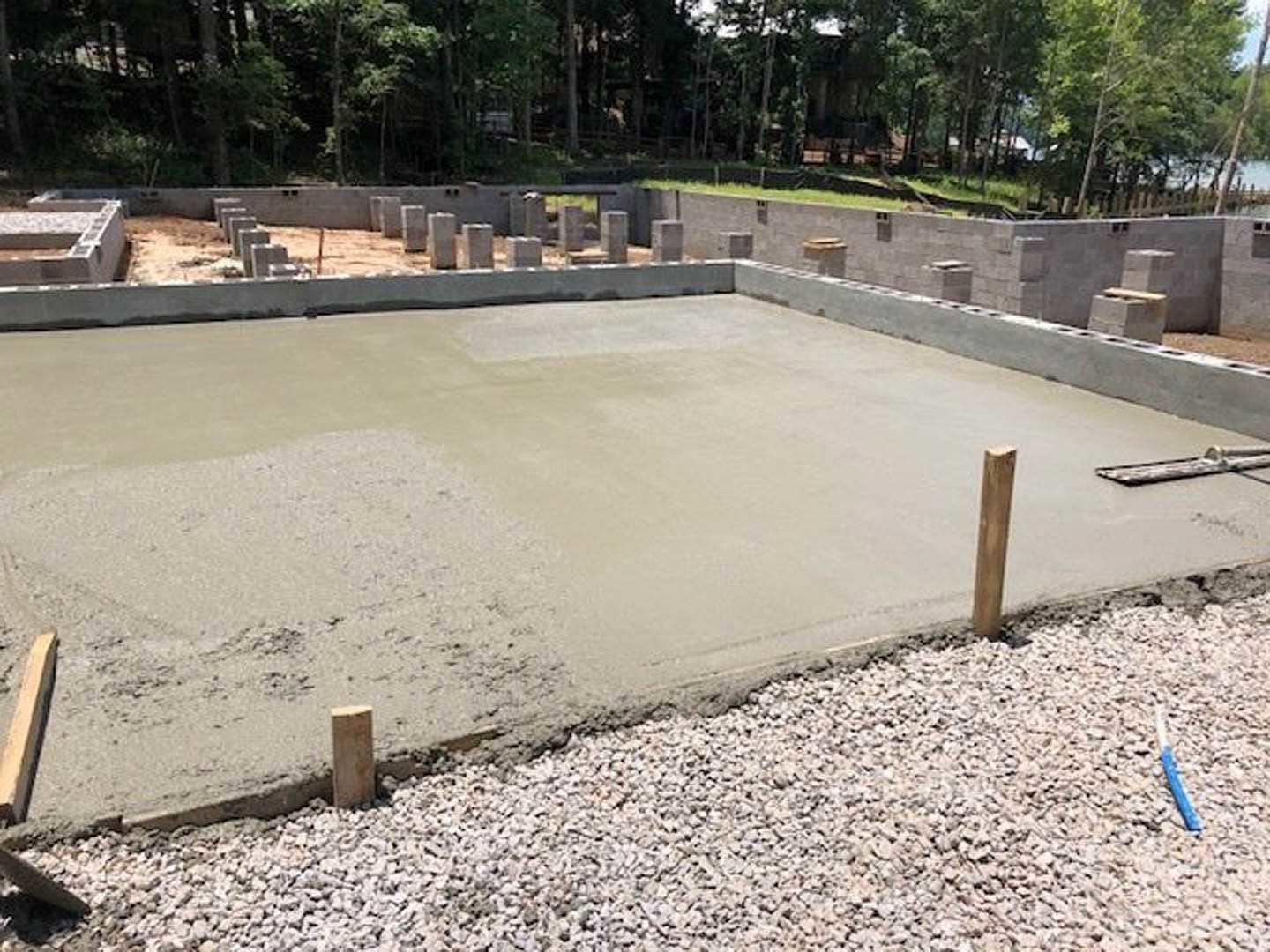 Freshly poured concrete foundation slab surrounded by gravel, wooden posts, and a wire fence in a wooded outdoor setting.