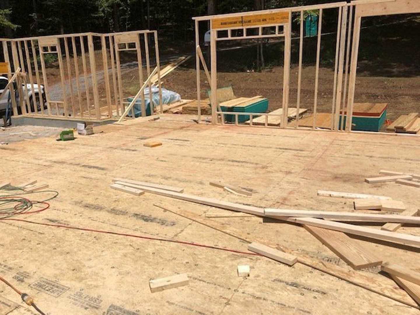 Wood-framed structure under construction with exposed lumber and concrete foundation, scattered wood planks and pieces on ground, white car parked in background, trees visible
